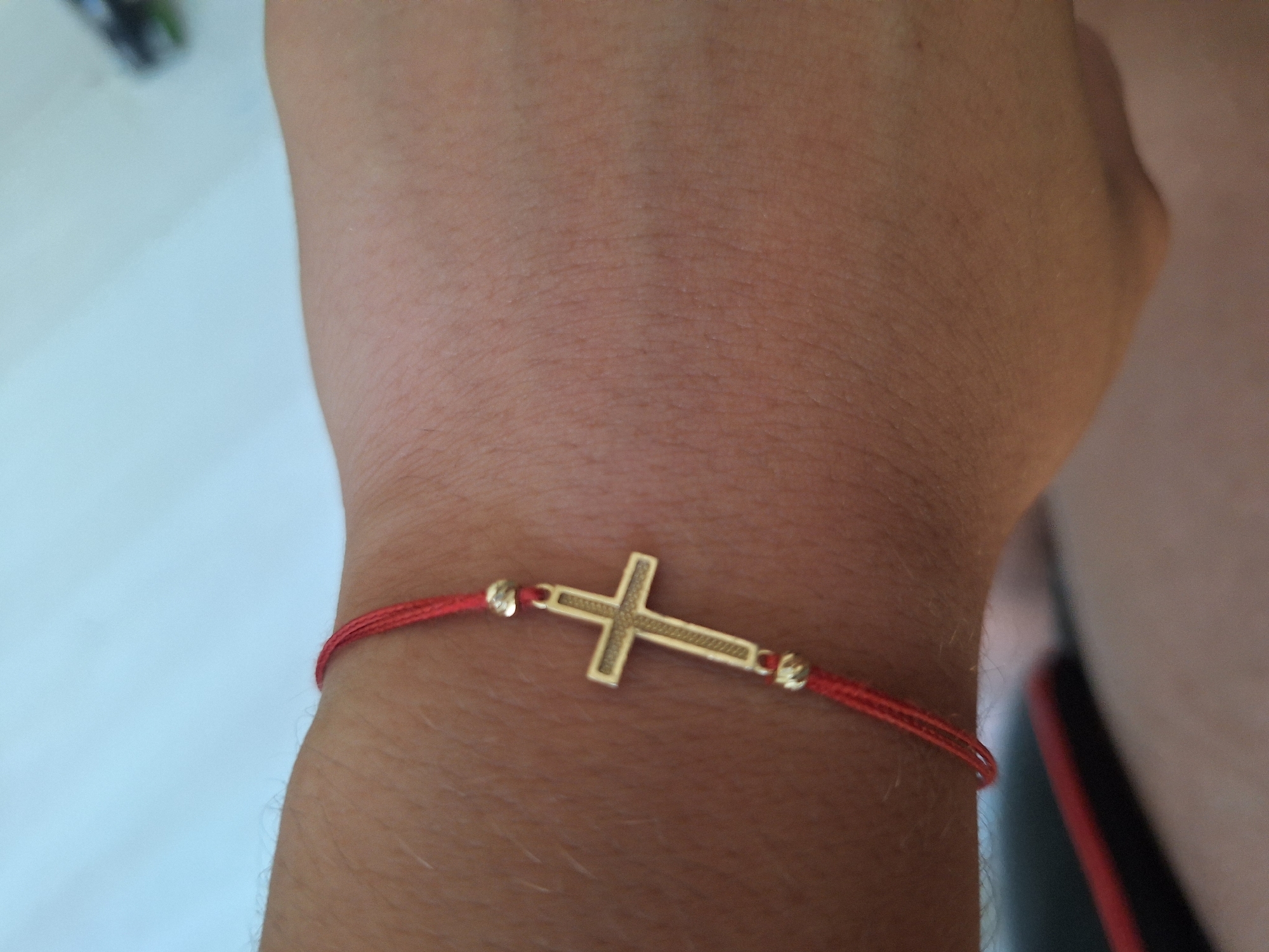 A close-up shot captures a red string bracelet with a gold cross charm and two small gold beads on a person's wrist. The bracelet is positioned on the underside of the wrist, with the cross facing outward. The skin appears tan and slightly textured, with visible hair follicles and fine lines. The background is a blurred light blue, suggesting an indoor setting. The image does not contain any information or visual cues to indicate the city of Varna, Bulgaria.