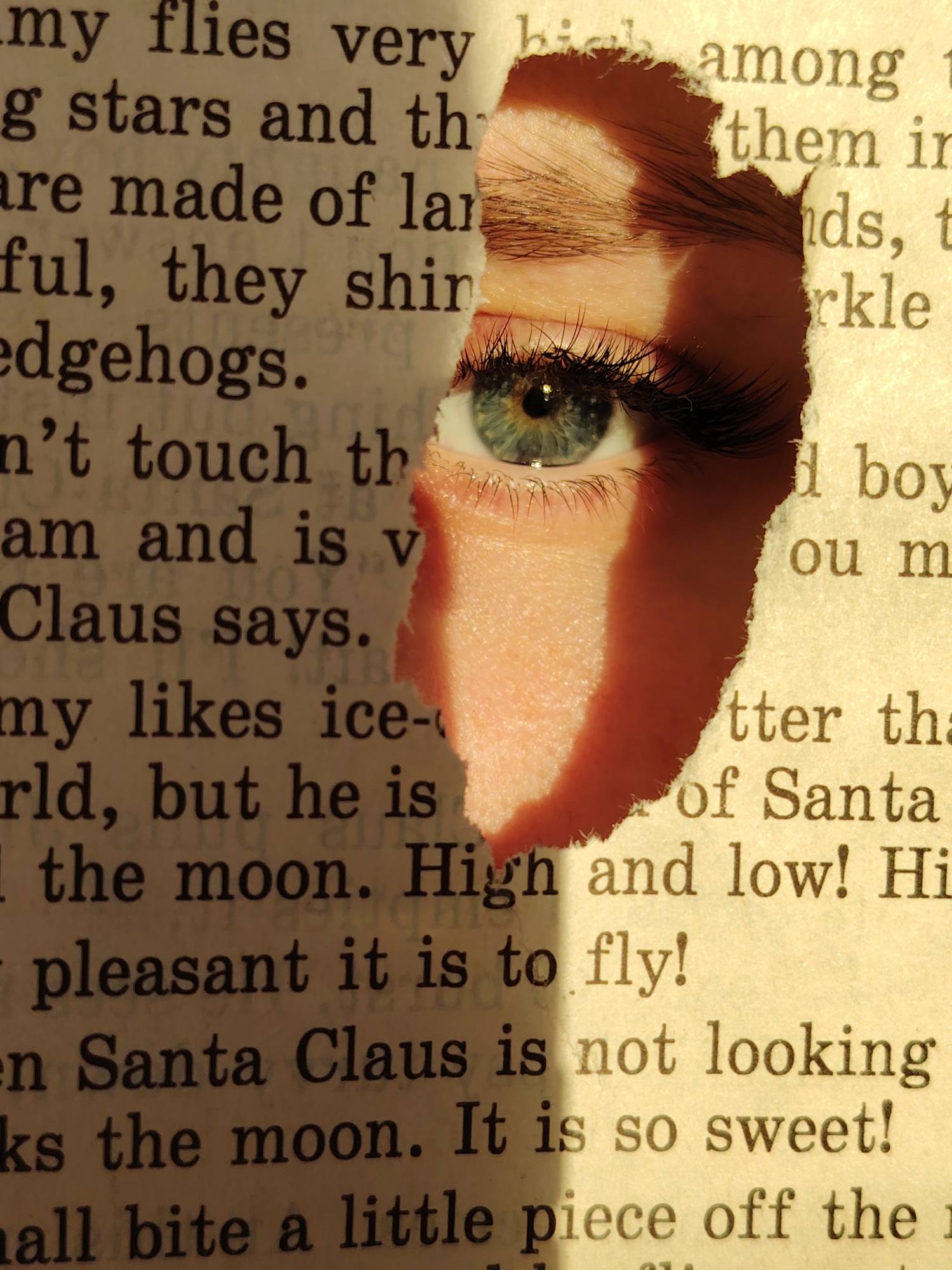 The image features an eye peeking through a torn page of a book. The eye is light blue-green and has long eyelashes. The skin around the eye is fair. The book's page is yellowed and contains black text, phrases include "my flies very high among," "stars and th," "them in," and "Santa Claus is not looking." The overall lighting is warm. The location is not identifiable as Chaikovsky, Russia, from the image alone.