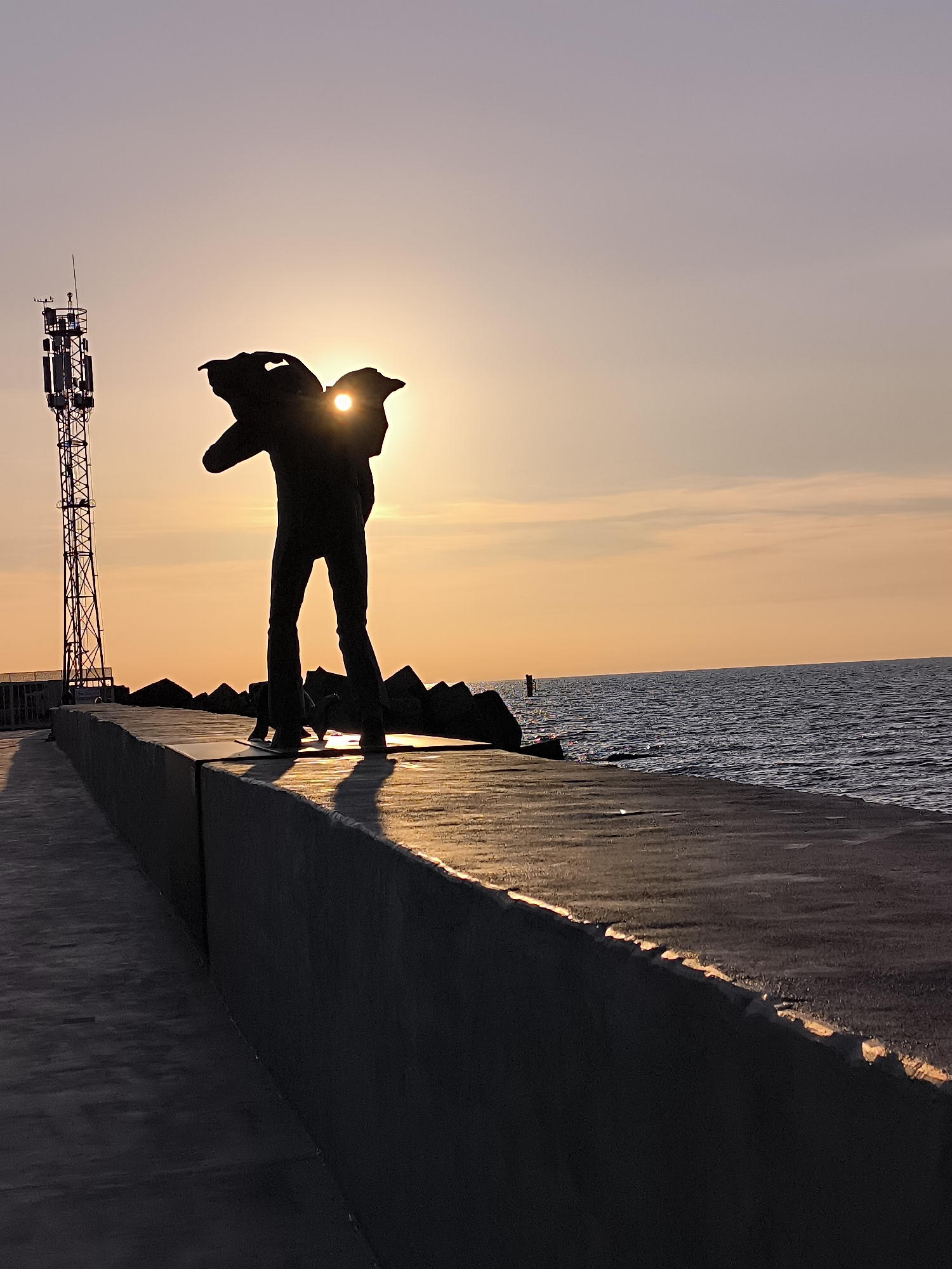 This media file depicts a coastal scene in Klaipėda, Lithuania, at either sunrise or sunset. The central subject is the dramatic silhouette of a person, likely a statue, standing on a concrete sea wall or pier. The figure is positioned with its back to the camera, facing towards the left side of the frame, and the bright sun is directly visible between its raised or outstretched hands, creating a striking halo effect.
To the left of the figure, a tall, multi-tiered metal communication tower equipped with various antennas stands against the sky. The sea stretches across the right side of the image, displaying gentle waves and reflecting the warm light. Behind the silhouetted figure and extending into the water, dark, jagged rocks form a breakwater.
The time of day is clearly indicated by the low angle of the sun and the warm, vibrant colors of the sky, which transitions from bright orange and yellow near the horizon to softer pinks and light purples higher up. Long shadows are cast by the figure and elements on the sea wall, further emphasizing the low light. The concrete surface of the sea wall in the foreground is partially illuminated, showcasing its texture. The overall atmosphere is serene and impactful, highlighting the contrast between the dark silhouettes and the brilliant sky and sun. No visible text is present in the image.