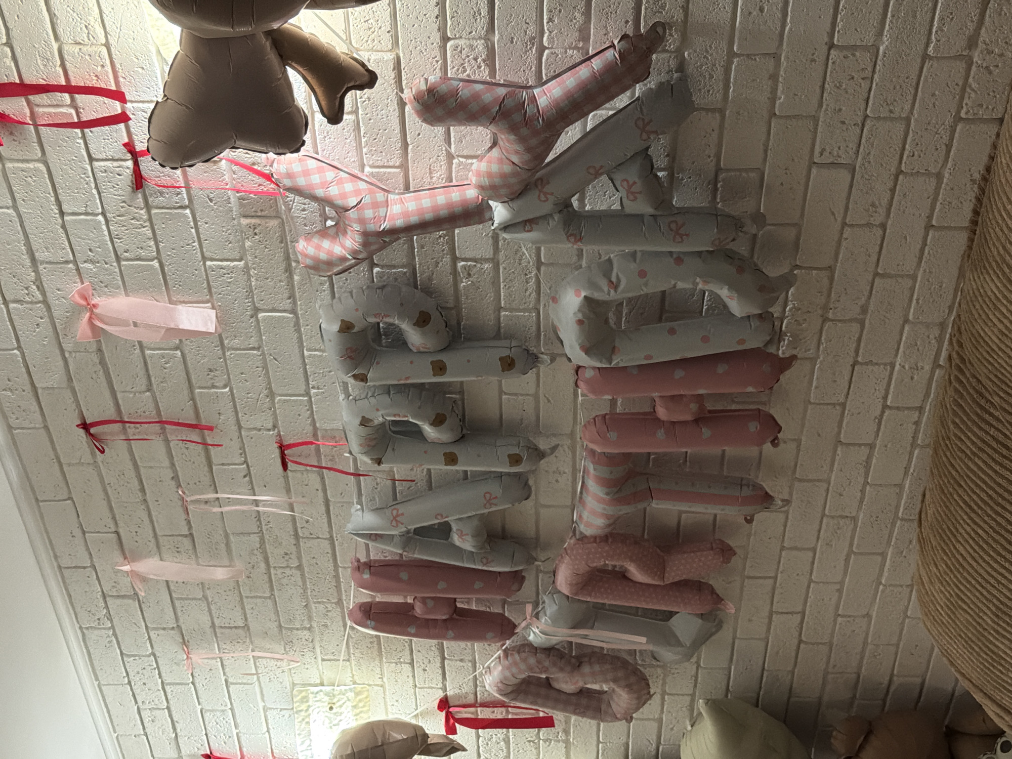 The image depicts a wall decorated with "Happy Birthday" balloons and ribbons. The balloons are shaped like letters and are patterned with small bows and bears on a light blue background, and pink gingham and polka dots on a pink background. Pink and red ribbons are tied to the wall with red bows, adding to the festive atmosphere. The wall itself appears to be made of white bricks. The scene suggests a birthday celebration is taking place indoors. There are no people visible in the image.