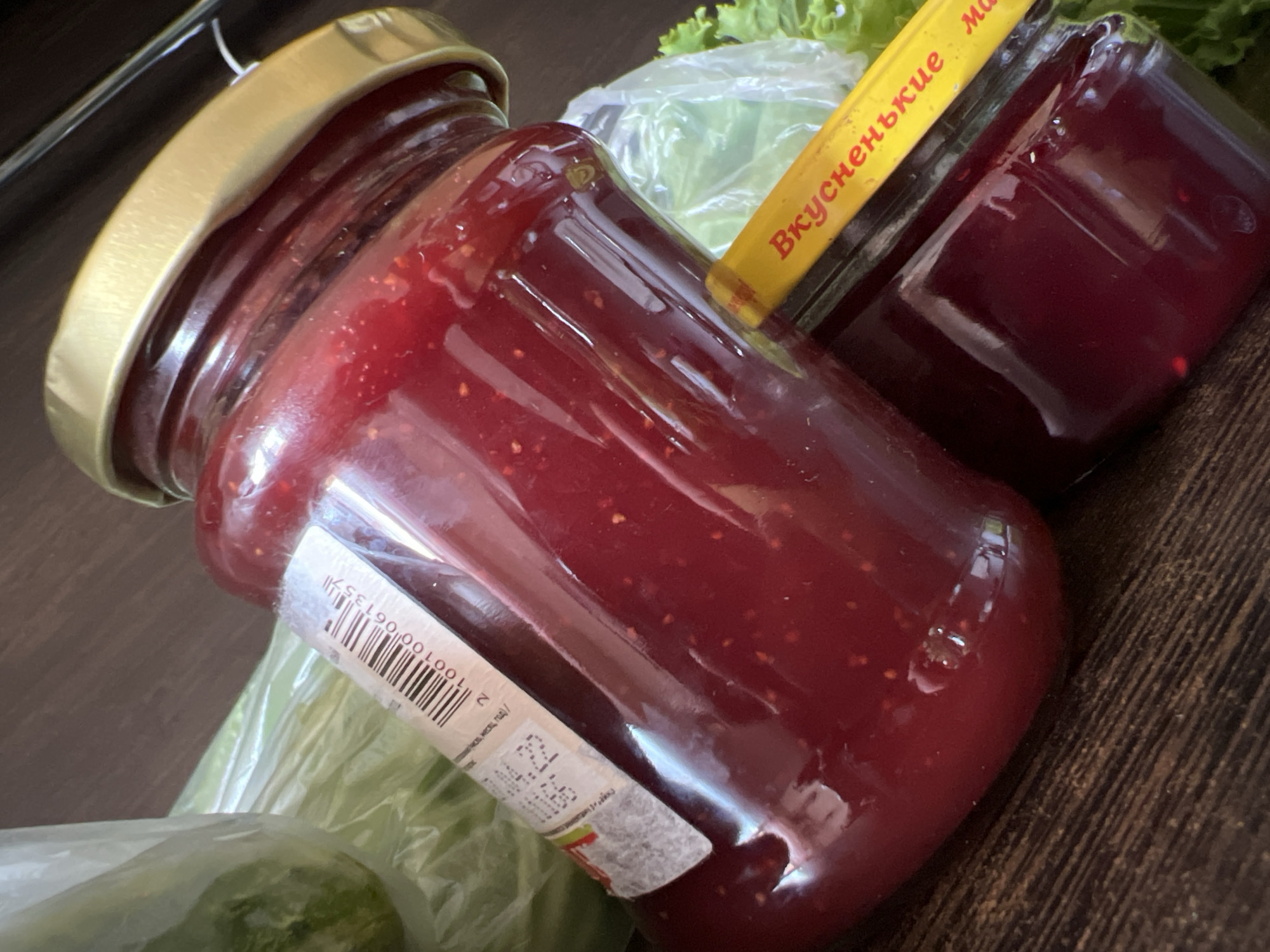 The image shows two jars of jam on a wooden surface. The larger jar is in the foreground and is mostly filled with a dark red jam. The lid of the larger jar is gold colored. A label on the larger jar has a barcode and some text. A smaller jar of jam is partially visible behind the larger jar, also filled with dark red jam. A yellow label with Russian text, "Вкусненькие ма", is affixed to the smaller jar. In the background, there is a green item in plastic wrapping, possibly vegetables. The lighting suggests an indoor setting. The scene is devoid of people or any activity. The location is Tolyatti, Russia.