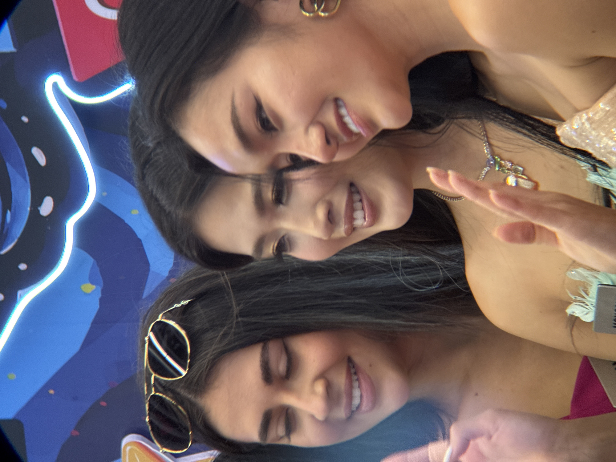 Three women are posing for a photo, with two of them looking at the camera and one looking to the side. The woman on the left is wearing sunglasses on her head and a pink top. The woman in the middle is wearing a necklace with colorful charms and a light blue and green dress. The woman on the right is wearing gold hoop earrings and a sparkly dress. They are standing in front of a blue backdrop with neon lights. The image appears to have been taken at an event in Nha Trang, Vietnam, likely an outdoor setting given the lighting.
