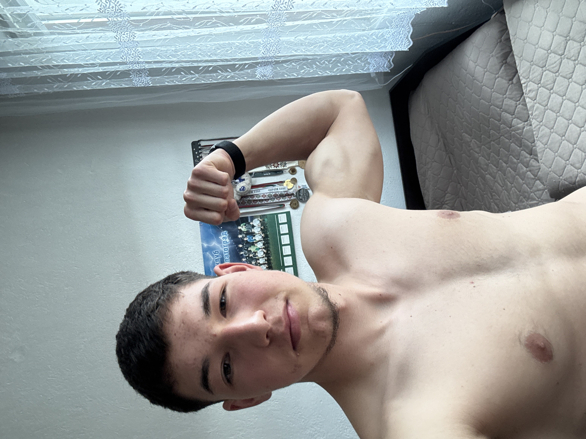 A shirtless young man with dark hair and a short beard poses with a flexed right bicep indoors. He is holding a calendar or poster featuring a soccer team photograph with writing in Cyrillic, possibly indicating it is from Zlatograd, Bulgaria. The background includes a wall, a window with a white sheer curtain, and a bed with a patterned gray comforter. He is wearing a black fitness tracker on his wrist.