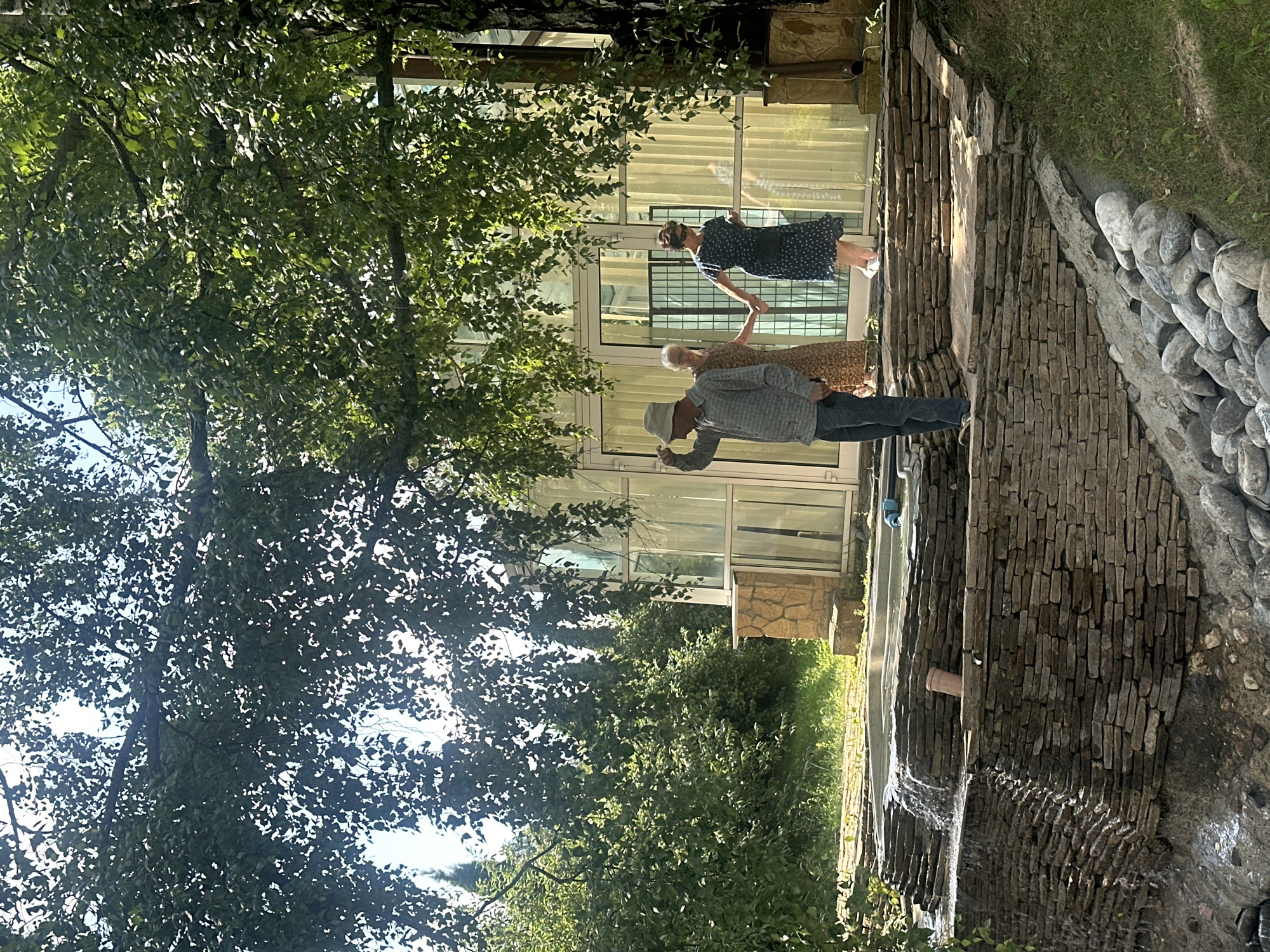 The image depicts three people standing outdoors in what appears to be a garden or a courtyard next to a modern building with large windows. The scene is oriented vertically, with the sky and treetops at the bottom and the ground at the top.
The subjects are two women and one man. The woman on the right is wearing a dark blue polka-dot dress and is holding hands with the woman in the middle, who is wearing a patterned dress. The man on the left is wearing a light-colored, checkered shirt and dark pants, and a light-colored hat. He is looking down and appears to be bowing or gesturing. The people seem to be engaged in some sort of interaction, possibly dancing or a ceremonial gesture, given their hand-holding and the man's pose.
The setting includes a building with a glass facade, a stone-paved pathway or terrace, and lush green trees and foliage. There is also a rock wall with water flowing down it, creating a small waterfall, and a grassy area to the right. The presence of flowing water suggests a possibly warm and sunny day, and the abundance of green suggests it is likely spring or summer. The lighting indicates it is daytime, with sunlight filtering through the leaves.
There is no visible text in the image. The overall mood seems pleasant and serene, with the natural elements of trees and water contributing to a tranquil atmosphere. The architecture of the building suggests a modern or perhaps a residential setting. The location context provided is Bogdanikha, Russia.