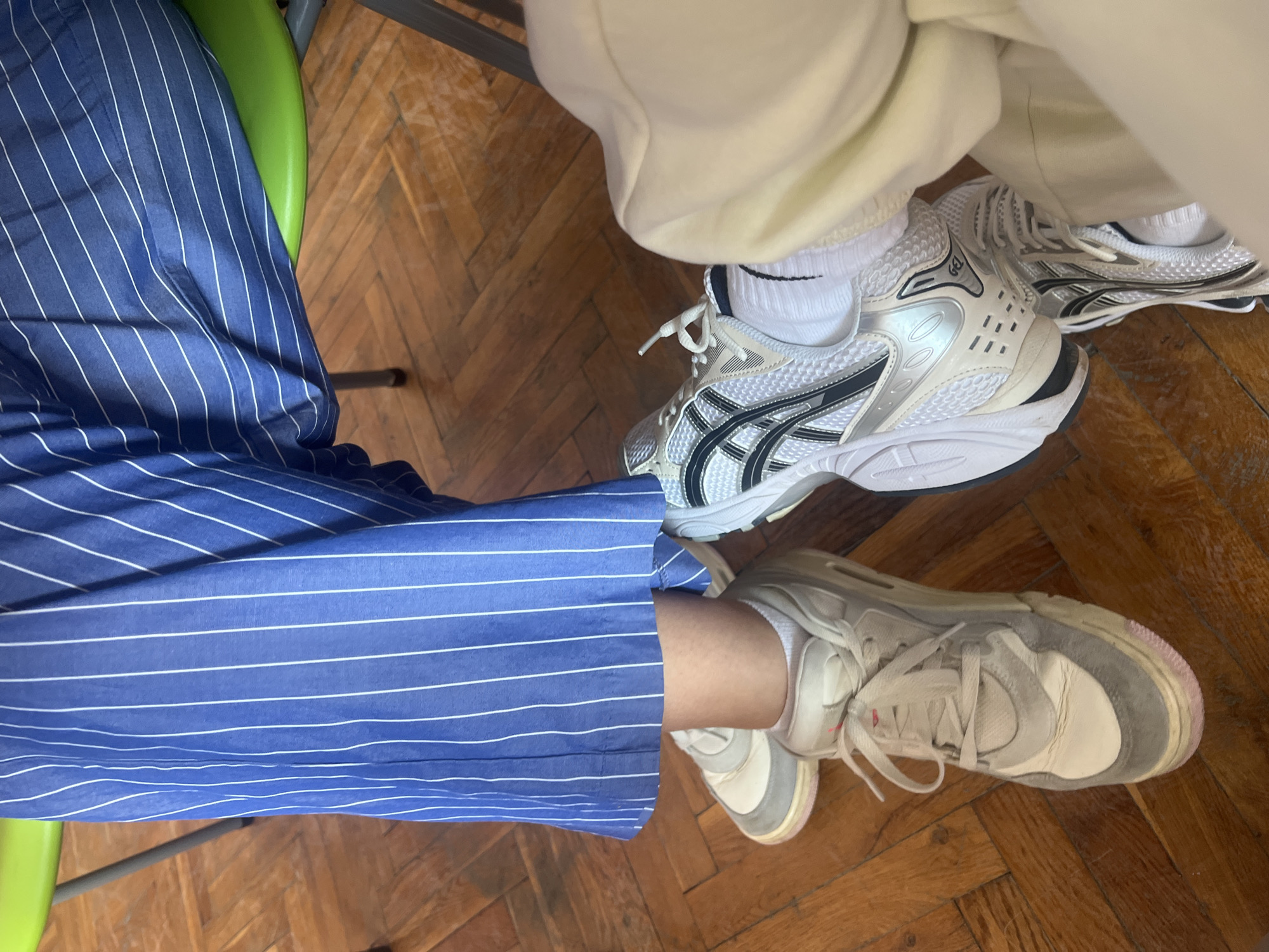 An overhead shot captures the lower halves of two individuals, seated indoors, with their legs and feet visible. The setting is characterized by a warm-toned wooden parquet floor, with hints of a green chair and dark furniture legs.
One person, positioned on the left, wears blue trousers adorned with thin white vertical stripes. Their leg is partially obscured by the fabric, and they are wearing a light-colored, possibly off-white, sneaker with grey and pink accents on the sole. White socks are visible above the shoe.
The second person, on the right, wears light, possibly cream-colored, casual trousers or sweatpants. Their feet are clad in white sneakers with prominent silver, black, and white detailing. The design suggests an athletic shoe style, with "GEL" text visible on the outer side of the left shoe, indicating an ASICS brand. White socks with a black Nike swoosh are seen above these sneakers.
Both individuals appear to be in a relaxed, casual environment, likely sitting on chairs, given the angle of the shot and the presence of chair parts. The overall impression is one of comfort and informality. No specific time of day or event is discernible, but the indoor setting and casual attire suggest a leisurely moment.