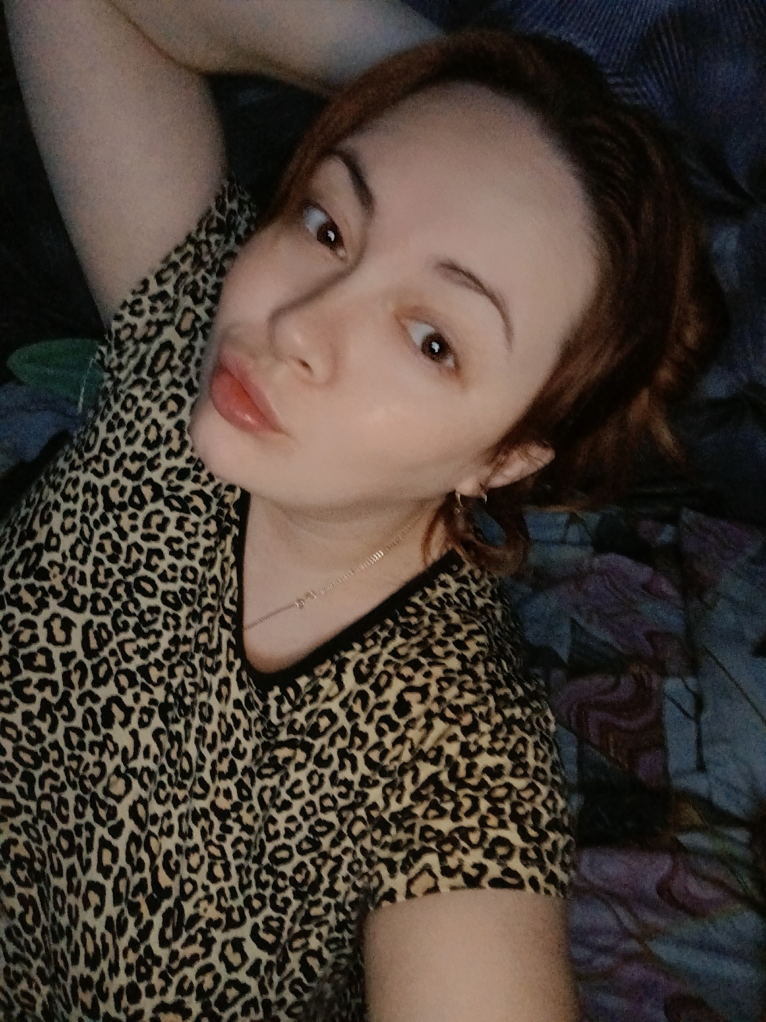A woman with reddish-brown hair is lying down, posing for a selfie. She is wearing a leopard-print top and a thin gold necklace. She has her lips pursed in a kissing gesture. The background is a dark blue and purple patterned surface. The location cannot be determined from the image.