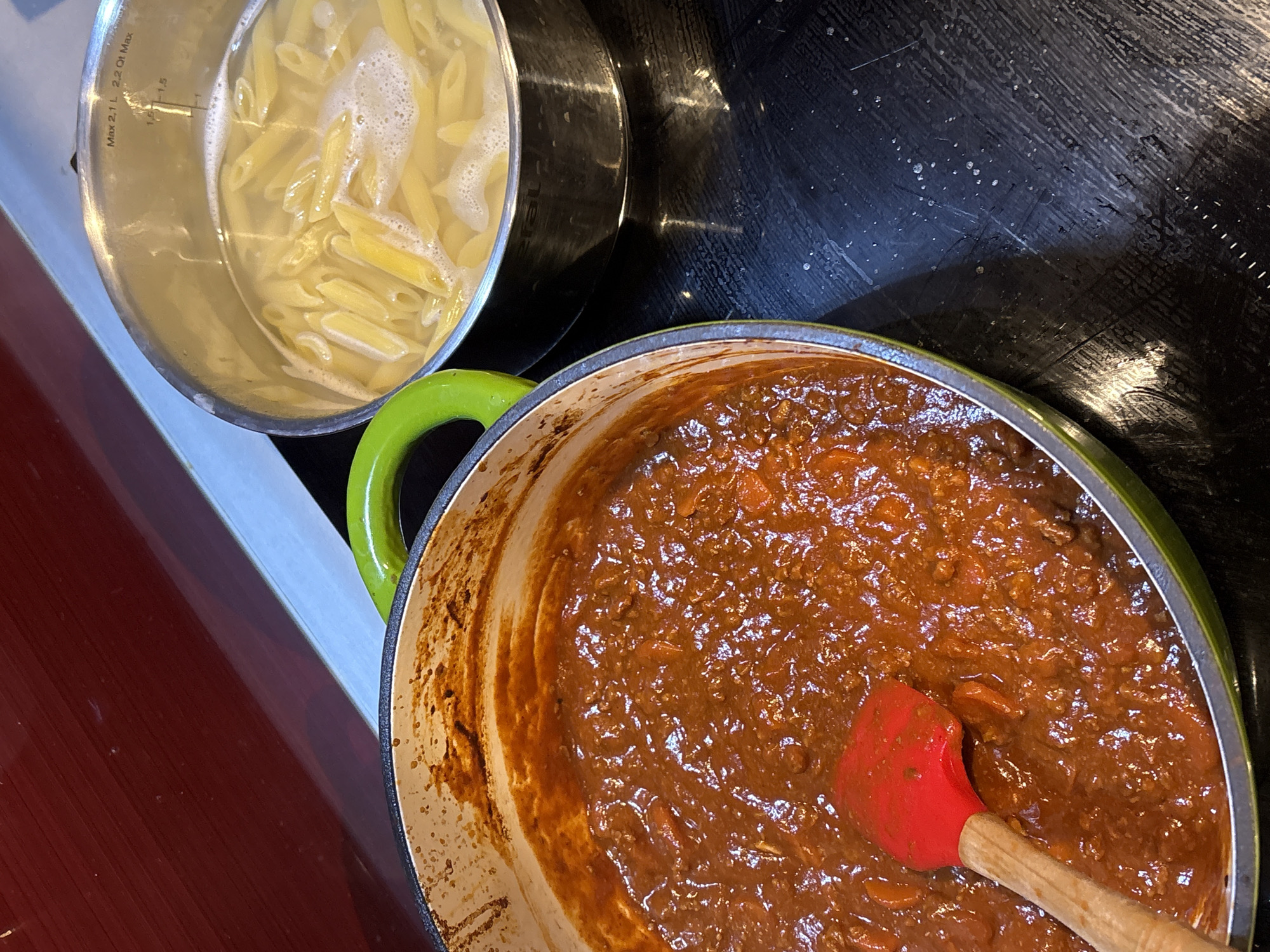 The scene captures a close-up of a kitchen countertop with two cooking pots on a dark, reflective electric or induction hob. On the left, a large, wide-mouthed pot with a light green exterior handle and rim contains a thick, dark reddish-brown sauce, appearing to be a ragu or bolognese, with visible chunks of meat or vegetables. A red silicone spatula with a light wooden handle rests partially submerged in the sauce. On the right, a stainless steel saucepan holds penne pasta boiling in foamy water. The saucepan features internal volume markings, including "Max 2.1 L / 2.2 Qt Max" and increments at 1.5 L and 1 L, with the brand "Tefal" partially visible. The background consists of a reflective dark red surface, likely a backsplash, above a light grey horizontal strip. The specific location within London, United Kingdom, is not ascertainable from the image.