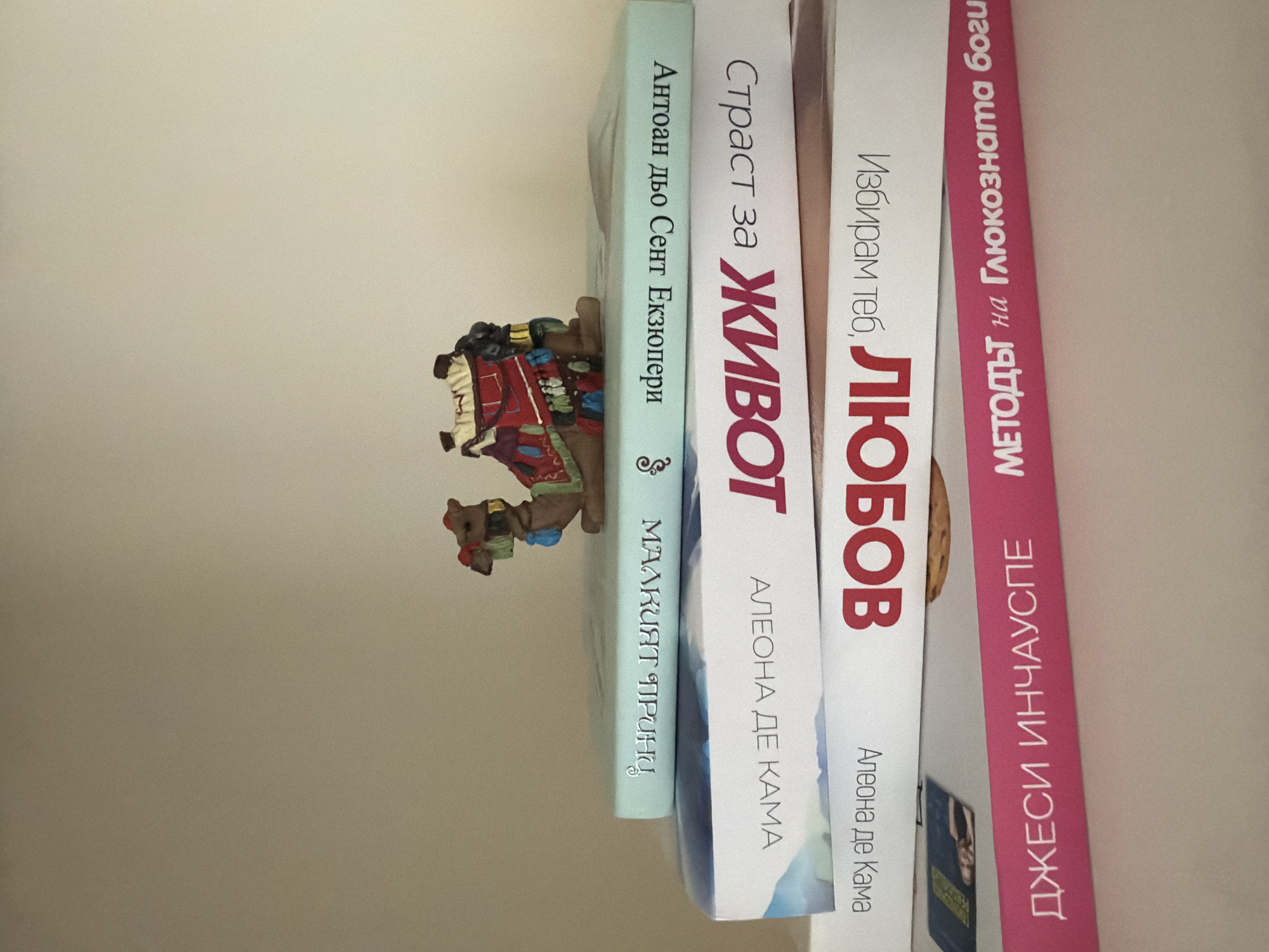 A small, decorative camel figurine with a colorful saddle is perched on top of a stack of four books. The camel is light brown with red, blue, and yellow accents on its saddle and a green saddlebag. It faces to the right. The books are stacked horizontally, with their spines facing upwards.
The top book has a light blue cover and features text in a silver, stylized font. The text reads "МАЛКИЯТ ПРИНЦ" and "Антоан дьо Сент Екзюпери".
The second book from the top has a white cover with blue watercolor-like artwork on the left side and text on the right. The text is in large, bold red letters and reads "ЖИВОТ" and "Страст за". Beneath that, in smaller black text, it says "АНЛЕОНА ДЕ КАМА".
The third book from the top also has a white cover with red text. The red text is in large, bold letters and reads "ЛЮБОВ" and "Избирам теб". Beneath that, in smaller black text, it says "АЛЕОНА ДЕ КАМА".
The bottom book has a bright pink cover with white text. The text reads "ДЖЕСИ ИНЧАУСПЕ" and "МЕТОДЪТ на Глюкозната богиня".
The background is a plain, light-colored wall. The scene is lit from the front, casting minimal shadows. There are no people or animals present other than the figurine. The image appears to be taken indoors, possibly on a shelf or table. The location is not specified in the image or any accompanying metadata.