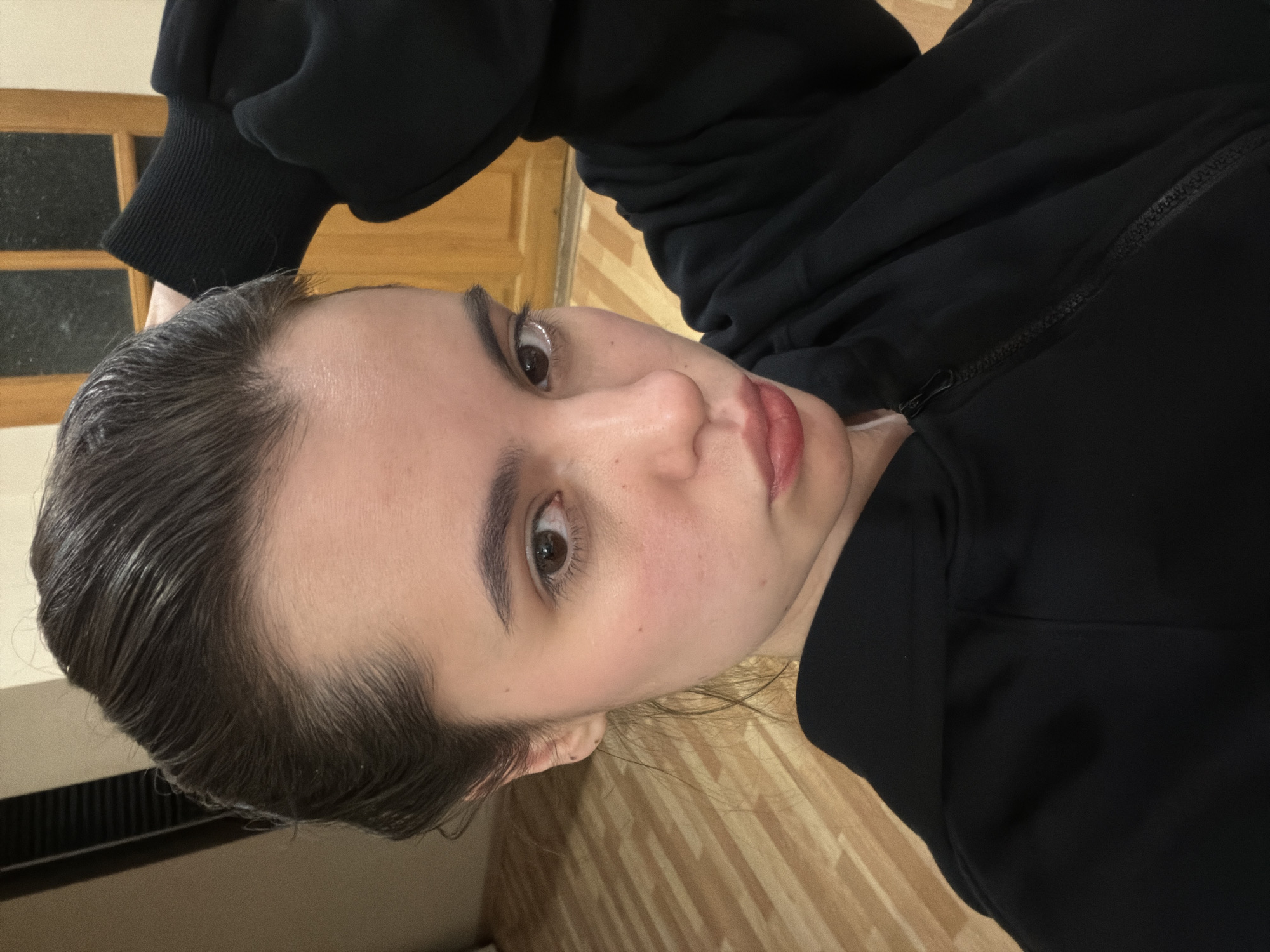 A person with fair skin and dark brown hair is seen in a close-up, eye-level shot. The hair is slicked back and has a wet appearance. The person is wearing a black hooded sweatshirt with the zipper partially visible. The background includes a wooden doorframe, a wooden floor, and what appears to be a radiator or air conditioning unit. The location is likely indoors. No specific landmarks identifying Perm, Russia are visible in the image.