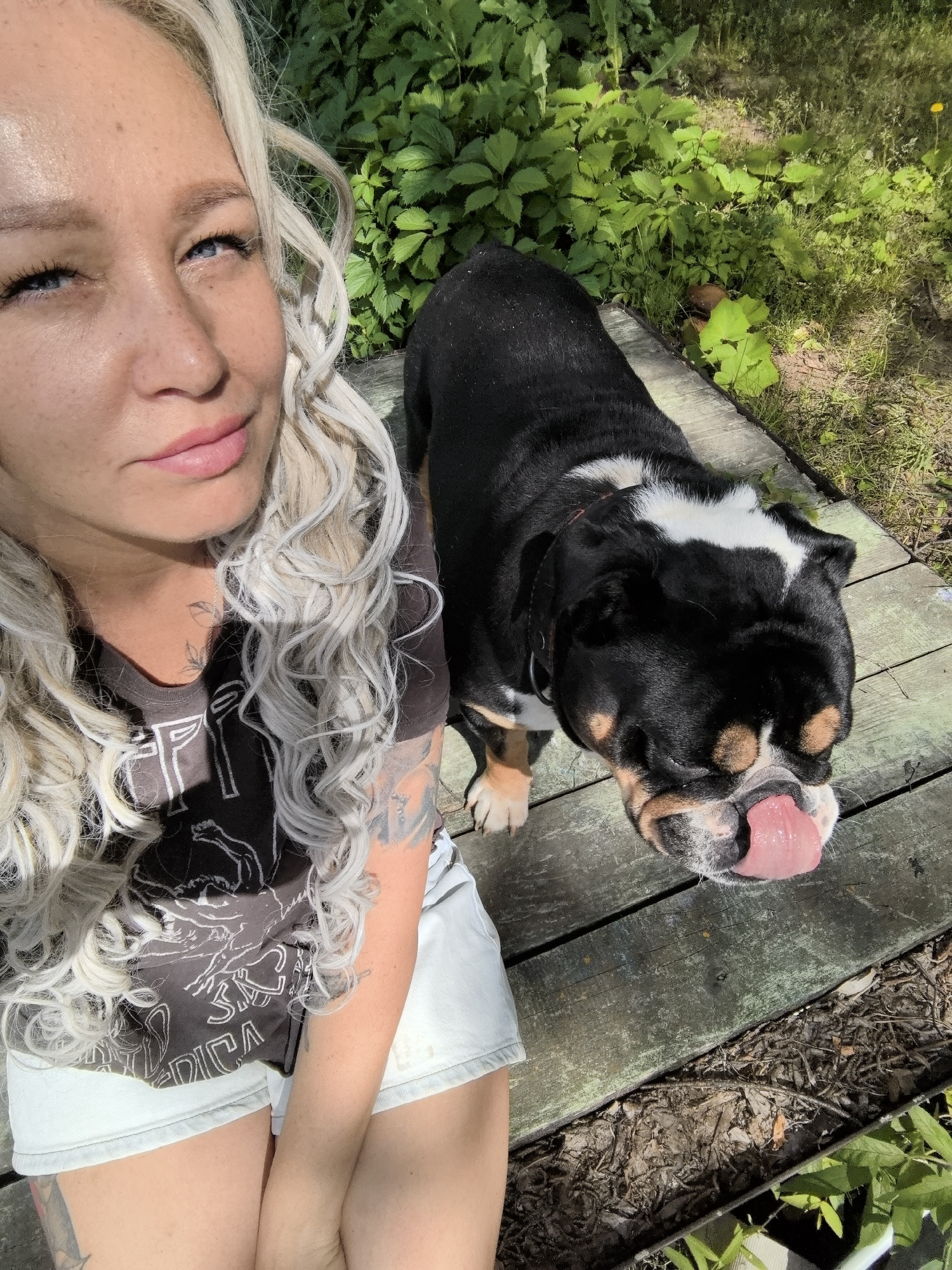In Noviy, Russia, a medium shot captures a Caucasian woman with long, wavy blonde hair, wearing a dark t-shirt and light-colored shorts. She is positioned on the left side of the frame, looking slightly upwards and to the right, with a partial view of her face and upper body. A black and white bulldog, with its tongue hanging out, is in the foreground on the right, facing the woman. Both are situated on a wooden deck. The background consists of lush green foliage under bright sunlight. The image appears to be a selfie taken outdoors.
