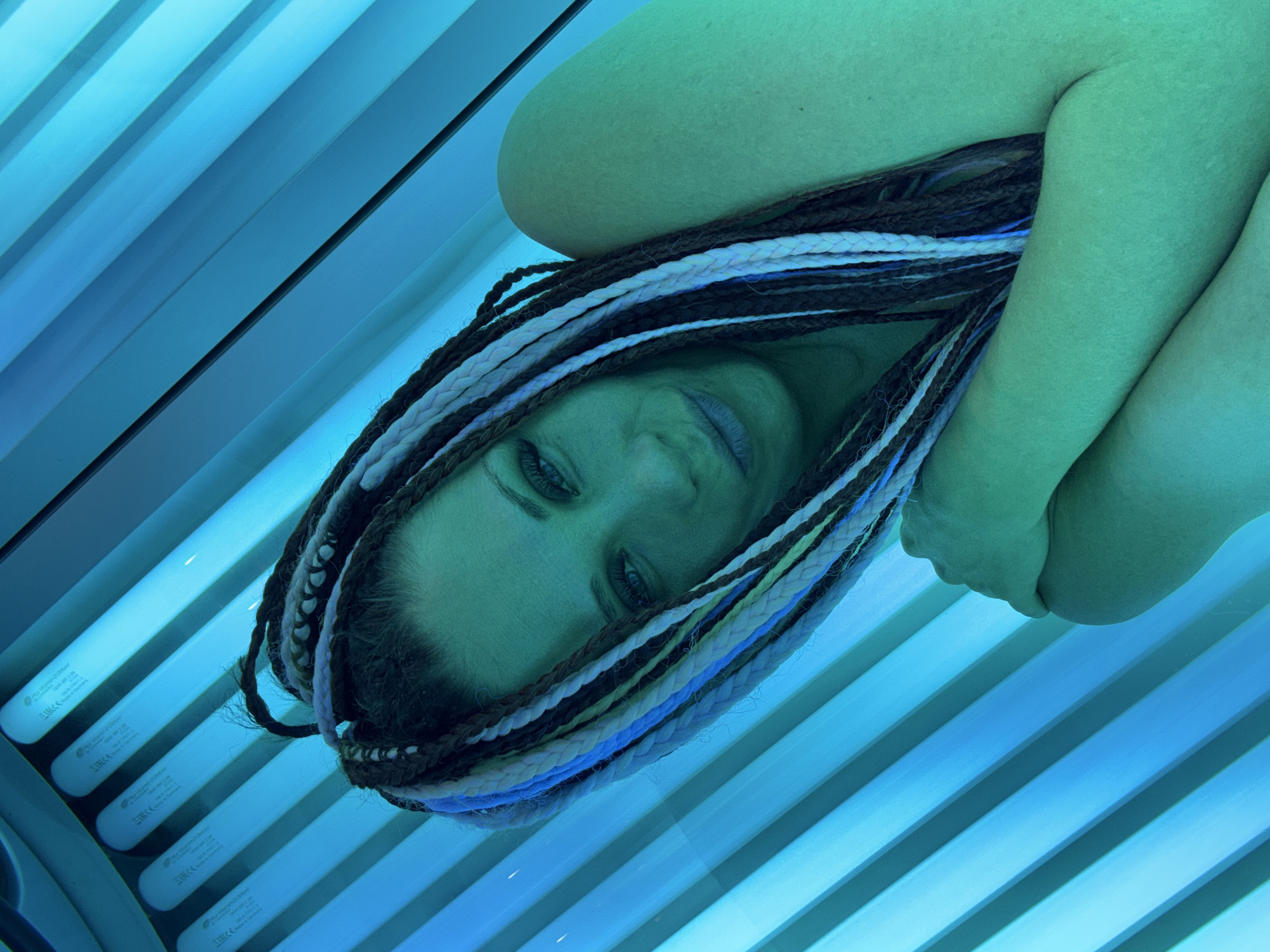 This image presents a high-angle, close-up shot of a person lying in a tanning bed, illuminated by its distinct blue light. The subject is positioned diagonally, with their head in the lower-left section and their body extending towards the upper right.
The central subject is a woman with long, multi-colored dreadlocks, which include shades of dark brown, light brown, white, and blue. Her face is turned slightly upwards, featuring a subtle, relaxed smile and blue eyes. Her skin, especially on her face and visible limbs, has a pronounced greenish-blue tint due to the intense blue light from the tanning lamps.
The background is dominated by the interior of the tanning bed, characterized by several parallel fluorescent tubes glowing with a bright blue light. These tubes line the left side of the frame. Visible text on the white casings of the tubes includes "RUVA Tanning & D-Luxx", "RUVA D-LUXX 120W", and "Made in Germany". Other identifiable symbols include the CE mark and a crossed-out wheeled bin logo, indicating product compliance and disposal instructions.
The scene depicts the activity of using a tanning bed in an indoor, artificially lit environment. The cool blue and green hues, resulting from the tanning lamps, create a distinct, almost ethereal ambiance around the subject. The overall impression is one of relaxation during a tanning session.