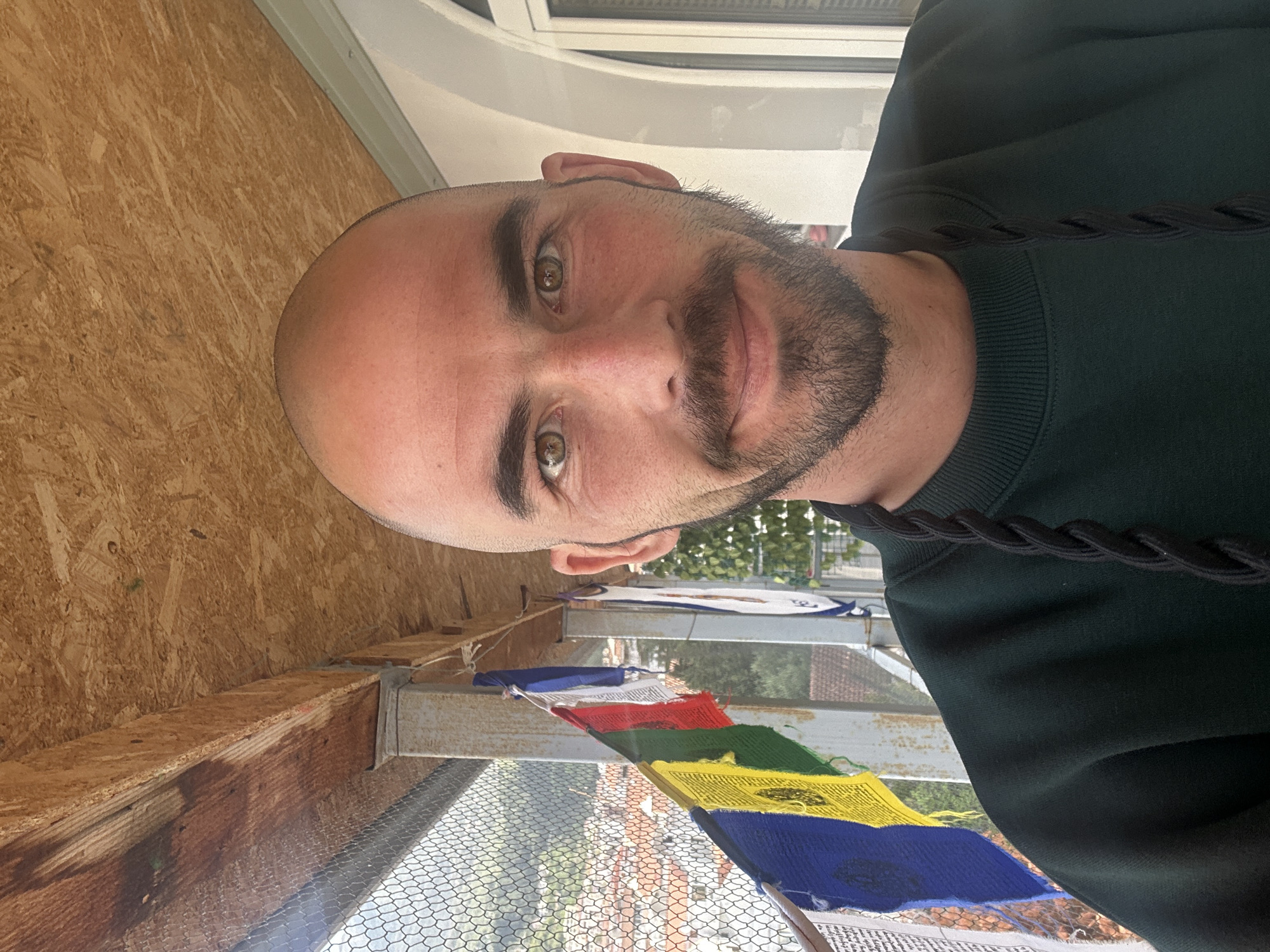 A bald man with light skin and a beard looks into the camera. He is wearing a dark green sweatshirt with a braided black strap. Behind him are colorful prayer flags attached to a metal structure covered with chicken wire. In the background are buildings, trees, and possibly houses, suggesting a residential area. The interior wood paneling suggests a sheltered or enclosed space. While no elements definitively place the image in Budva, Montenegro, the style of construction and vegetation are consistent with that region.