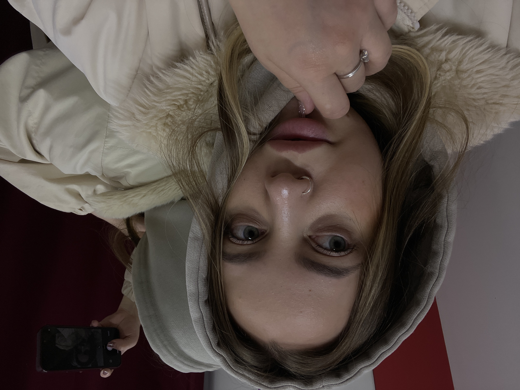 A young woman is holding a smartphone in her left hand and is pointing a finger from her right hand at her lips. She is wearing a beige hooded jacket with a faux fur trim. She has light brown hair and a silver nose ring. The background is divided between a red and white wall. The location is likely indoors. It is impossible to verify the location is Novopokrovskaya, Russia from the image alone.