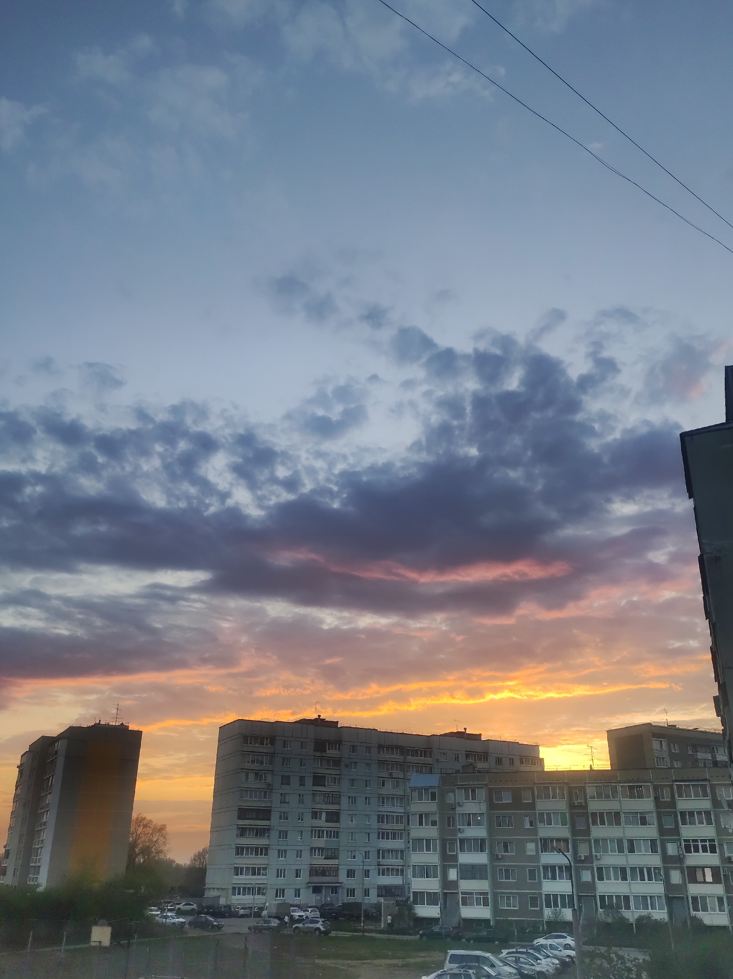 The image shows an urban sunset in Chaikovsky, Russia. Several multi-story apartment buildings dominate the view. A vibrant orange and yellow sunset illuminates the scene, with the sky above containing dark, textured clouds. Some electrical wires are visible in the upper right of the frame. Cars are parked in front of the buildings.