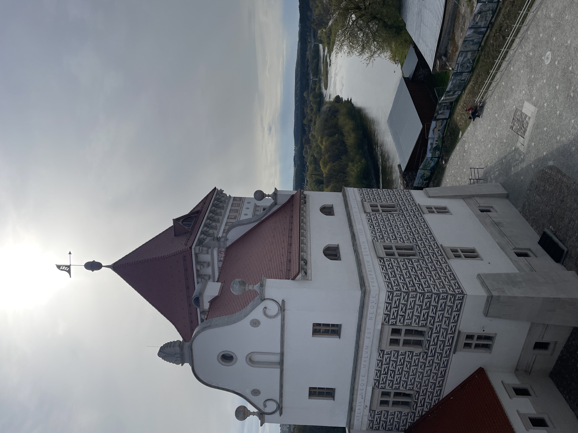 The image presents a high-angle view of a prominent historic building, likely a castle or palace, situated overlooking a river and surrounding landscape. The central subject is a multi-story white edifice, crowned with a distinctive red-tiled hip roof. Its facade is adorned with ornate architectural details, including decorative gables, scrolled elements, and prominent sgraffito-like patterns of repeating dark geometric shapes on its lower sections. A weather vane marked "1587" is visible on the building's main spire, silhouetted against a bright patch of sky. A Latin inscription runs along a band on the building's wall, stating: "MATERIA NEQVE MALVM EST. NEQVE PROPRIUM BONVM. SED ALIUUD."
From the elevated perspective, a wide river meanders through a lush, green landscape of trees and foliage. A bridge is faintly visible in the distance across the water. Adjacent to the building, a paved courtyard with patterned stones features an outdoor exhibit or display panels lining a fence. One individual, wearing a dark jacket, is seated on a bench in this area. The sky is largely overcast with light grey clouds, although a bright glare from the sun is visible towards the top left. The scene captures a static moment, highlighting the architectural grandeur of the building and its scenic natural surroundings, consistent with a historical site in Hrodna, Belarus.
