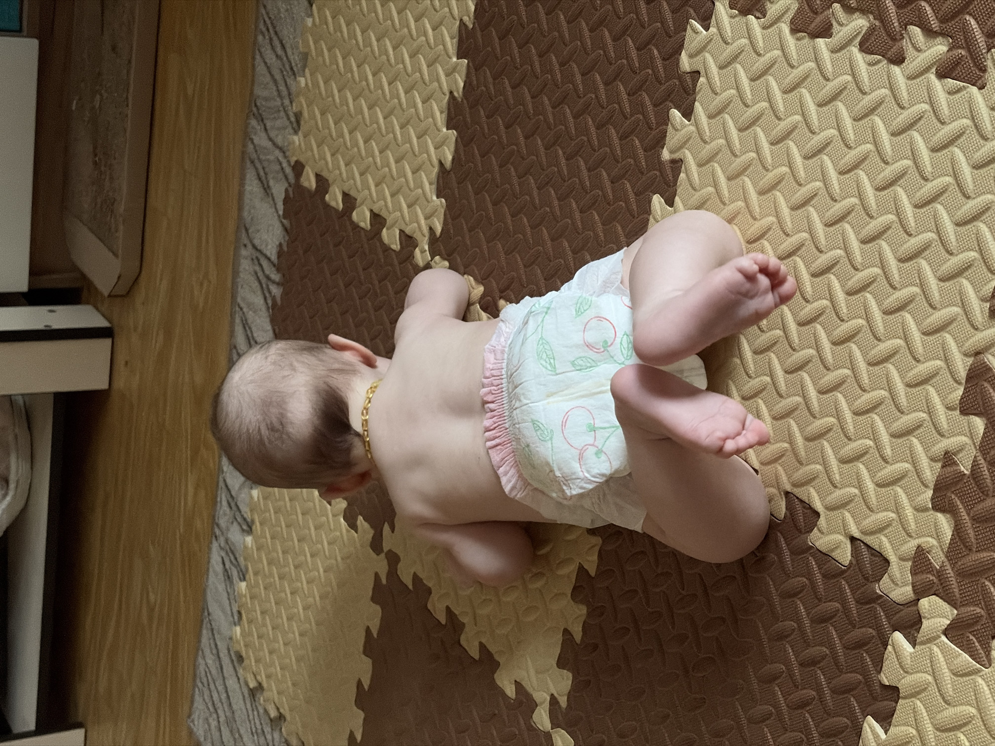 A baby lies on a brown and tan interlocking foam mat. The baby is wearing a diaper with a cherry print and a gold chain around their neck. The scene is indoors, next to a wooden cabinet. The location in the city of Chaikovsky, Russia is not discernible from the image.