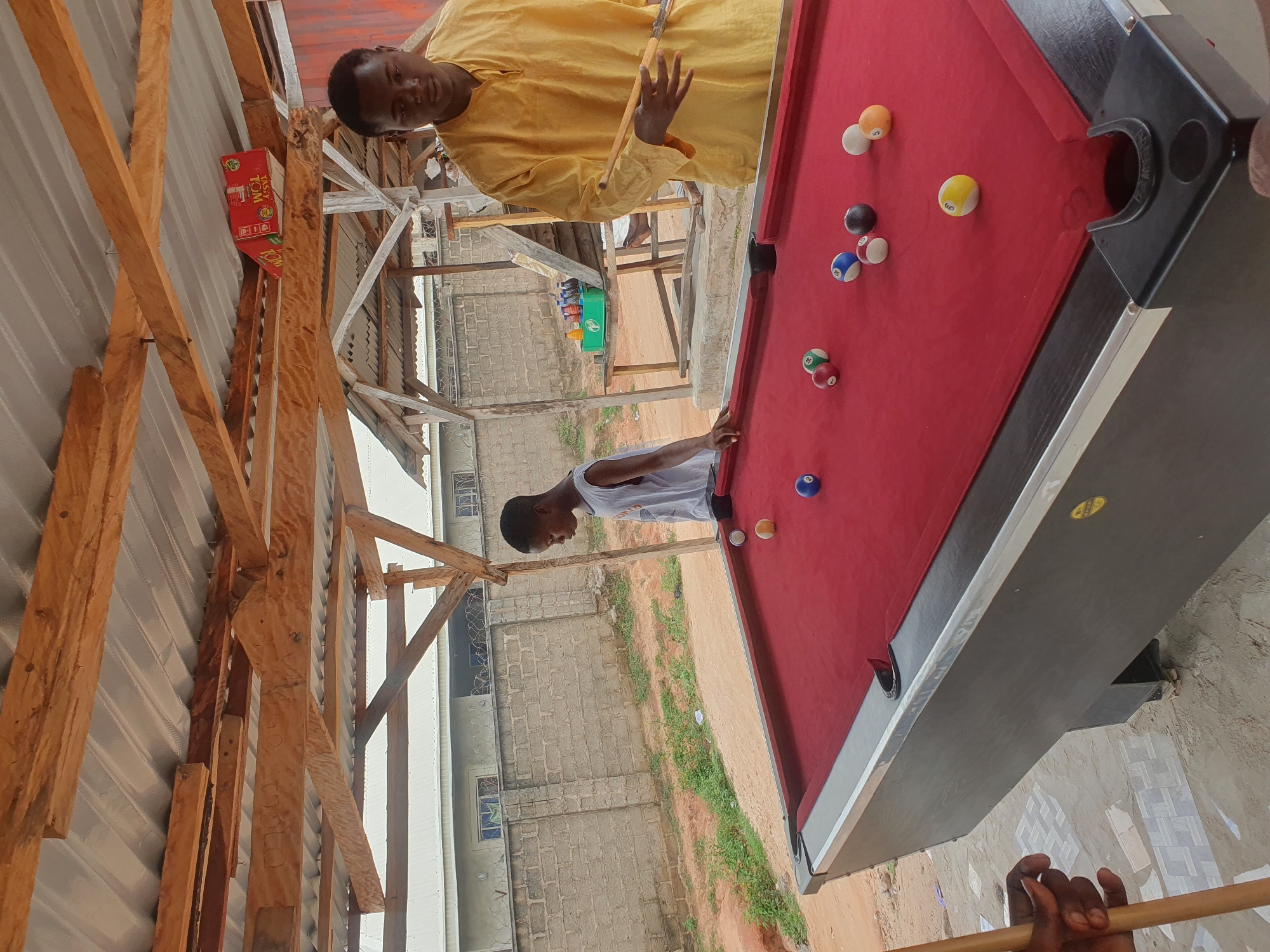 This is a medium shot of two young males at an outdoor pool table in Lokoja, Nigeria. The pool table has a red felt surface with several balls scattered across it, including a blue 8-ball, a yellow 9-ball, and an orange 5-ball. The table is made of dark wood with metal trim and black pockets.
The male on the left, wearing a light blue sleeveless shirt, stands by the pool table, his left arm resting on the edge. He appears to be observing the game. The male on the right, wearing a yellow tunic, holds two pool cues in his right hand, poised to strike a ball. He is looking down at the table, concentrating.
The scene is set under a rustic, open-air shelter constructed with corrugated metal roofing and wooden beams. In the background, a light brick wall separates the area from buildings with barred windows. The ground is a mix of sand and some scattered debris. To the right, a wooden table holds several bottles of drinks in a green crate. The overall impression is one of a casual, community gathering space.