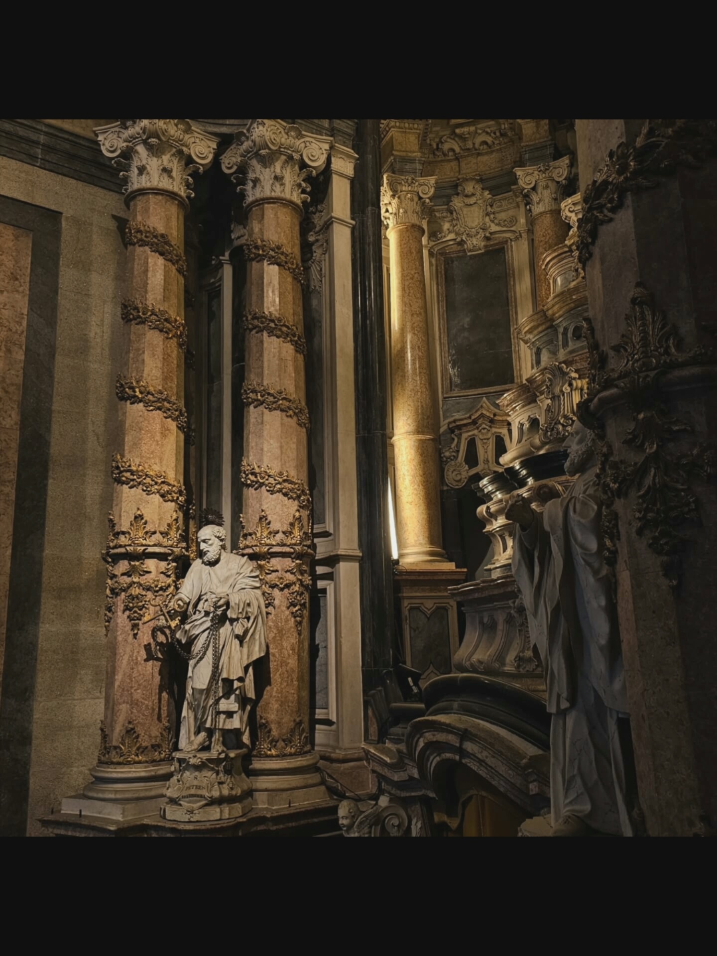 The image depicts the interior of a church, likely a basilica, with ornate Baroque architecture. Two marble statues of robed male figures are prominently featured. The statue on the left, positioned in the foreground, is of a bearded man holding a key and a chain, standing on a pedestal inscribed with Latin text. This is identified as Saint Peter. The statue on the right, further back and partially obscured, also appears to be of a similar religious figure, with hands clasped in prayer.
The statues are situated between massive, fluted columns adorned with gilded floral motifs and intricate capitals. The columns appear to be made of marble with reddish and beige veining. Behind the statues, the church's interior reveals more elaborate architectural elements, including gilded sculptures, niches, and arched structures. The lighting is dim, with a strong directional light source illuminating the right side of the scene, casting shadows and highlighting the textures of the marble and gold ornamentation.
The environment suggests a place of worship, with its grandeur and religious iconography. The location is identified as the Basilica di San Benedetto in Norcia, Italy.