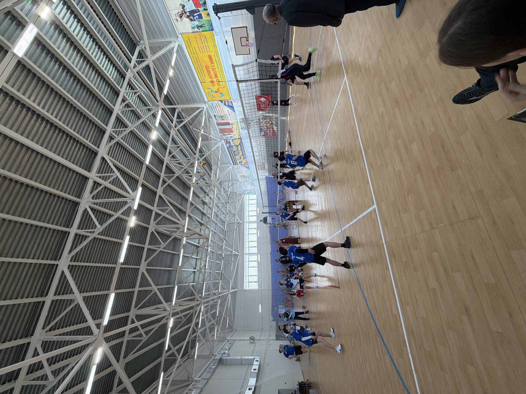 The image shows a volleyball game taking place in a gymnasium. The walls of the gymnasium are adorned with posters and banners. The game is actively in progress, with players from both teams positioned on their respective sides of the net, poised to hit the ball. Spectators are present, viewing the game from the sidelines. The gymnasium features a roof structure constructed of metal beams and supports. The location of the gymnasium is Bolshaya Murta, Russia.