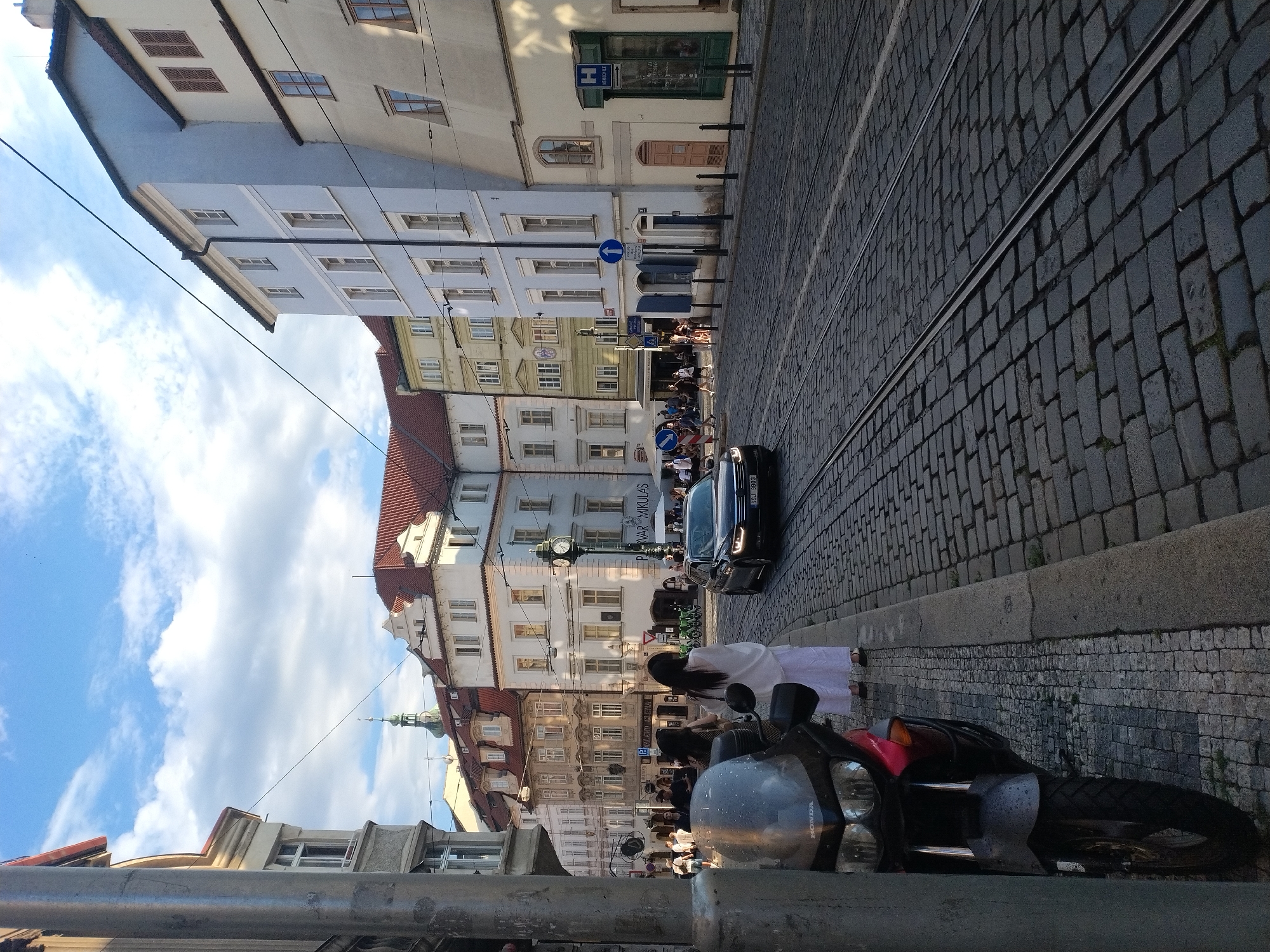 This image depicts a street scene in Prague, Czechia, likely during daylight hours, given the presence of shadows and the blue sky with clouds. The foreground is dominated by a dark-colored motorcycle parked on the left side of the cobblestone street. A woman wearing a white dress is standing on the sidewalk to the right of the motorcycle, facing away from the camera.
The street itself is paved with cobblestones, and tram tracks run down the center. In the middle ground, a black sedan is driving on the street. The buildings lining the street are varied in architectural style and color, with several stories and red-tiled roofs. Some buildings have visible windows and doors, and there are streetlights and signage visible. People are walking on the sidewalks and in the street, suggesting a moderately busy urban environment. The sky is a bright blue with scattered white clouds.
