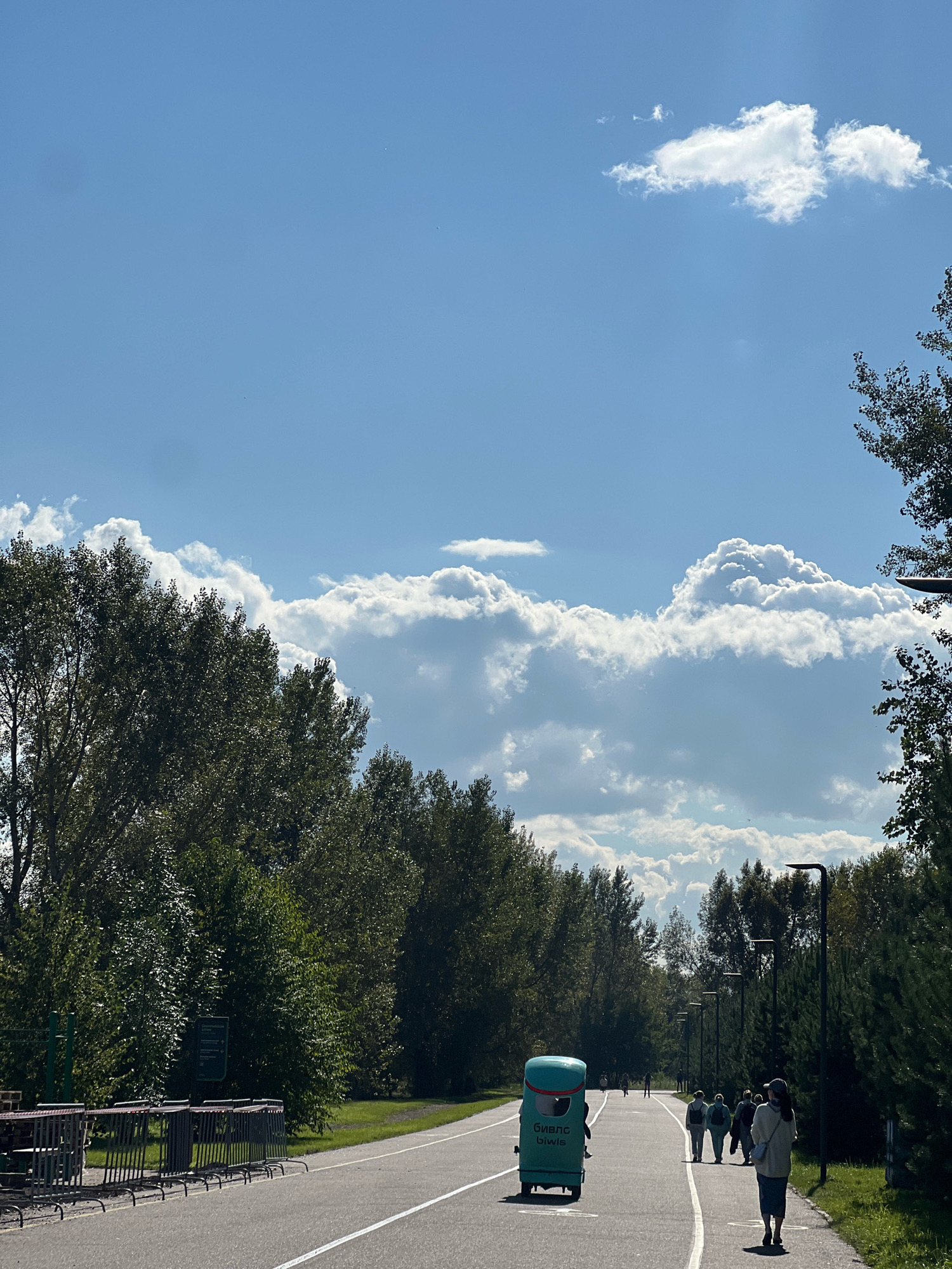 In a park on a sunny day with a bright blue sky and scattered white clouds, a paved pathway winds through a green landscape with numerous trees. On the pathway, a small, turquoise robotic vehicle, possibly a delivery or maintenance bot, is moving forward. Several people are walking or standing along the pathway, some appearing to be individuals or small groups. The scene is likely from a park in the city of Krasnoyarsk, Russia, given the presence of Cyrillic text on the robotic vehicle. The overall atmosphere is one of a pleasant, recreational outdoor setting.