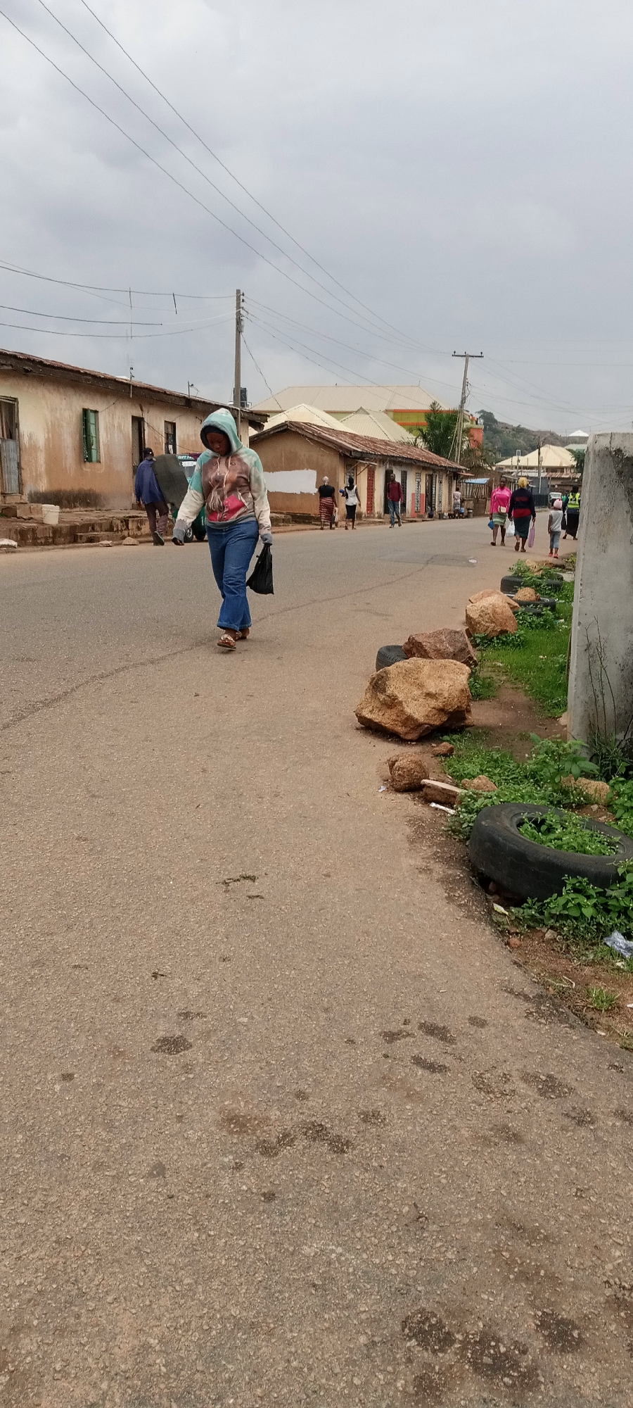 The scene depicts a street in Jos, Nigeria. In the foreground, a woman walks along the road, wearing a hooded sweatshirt, jeans, and sandals, carrying a black plastic bag. Several people walk in the background along the street and on the sidewalks in front of buildings. The buildings along the street are small, single-story structures with aged, weathered facades. Along the roadside in the foreground are rocks and a discarded tire with vegetation growing inside. The sky is overcast. Overhead, power lines stretch across the street.