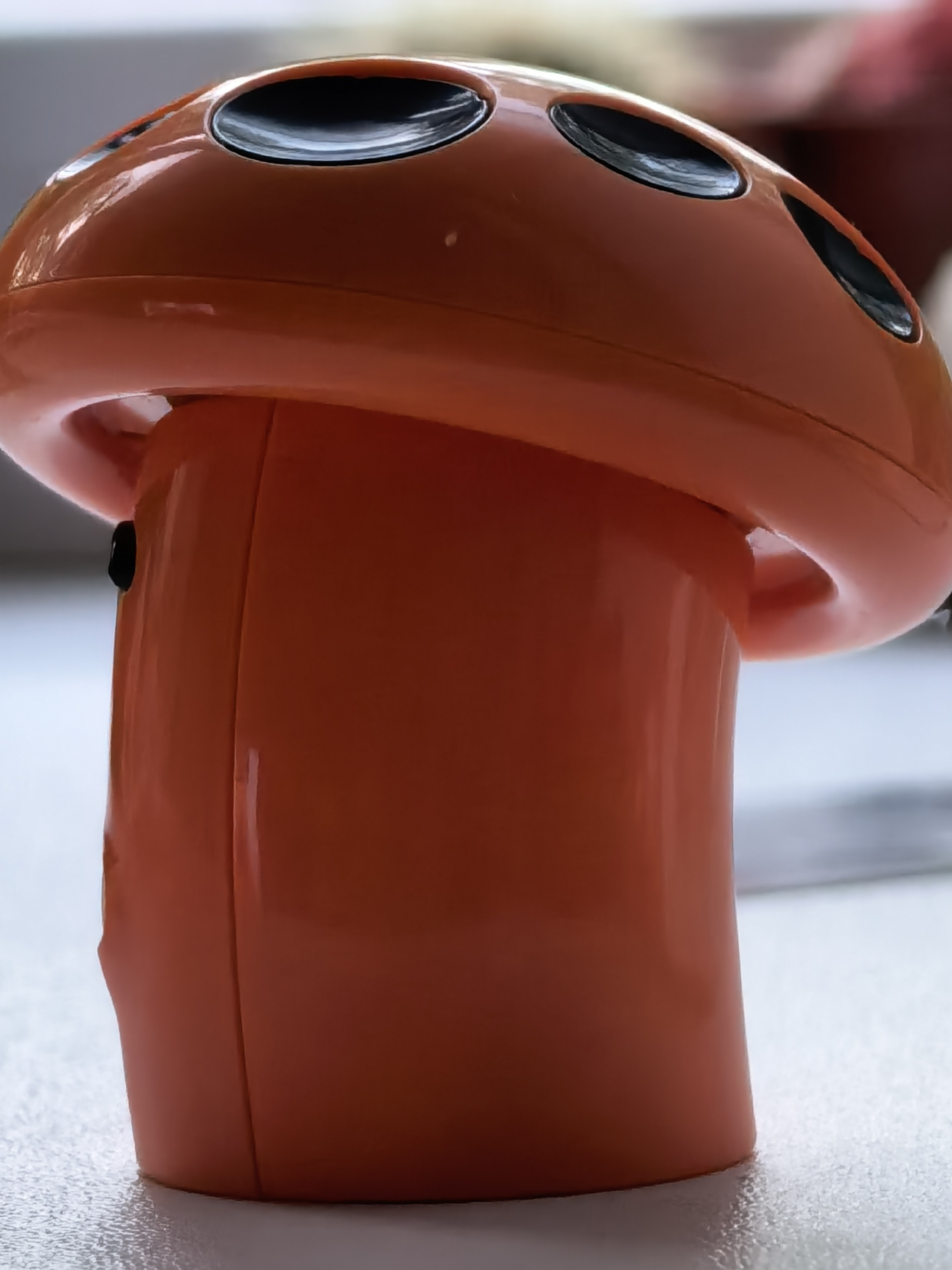 An orange, plastic, mushroom-shaped object with black holes on the cap sits on a light-colored surface. The object appears to be a child's toy or a decorative item. The surrounding environment is nondescript and does not reveal the location as Sterlitamak, Russia.