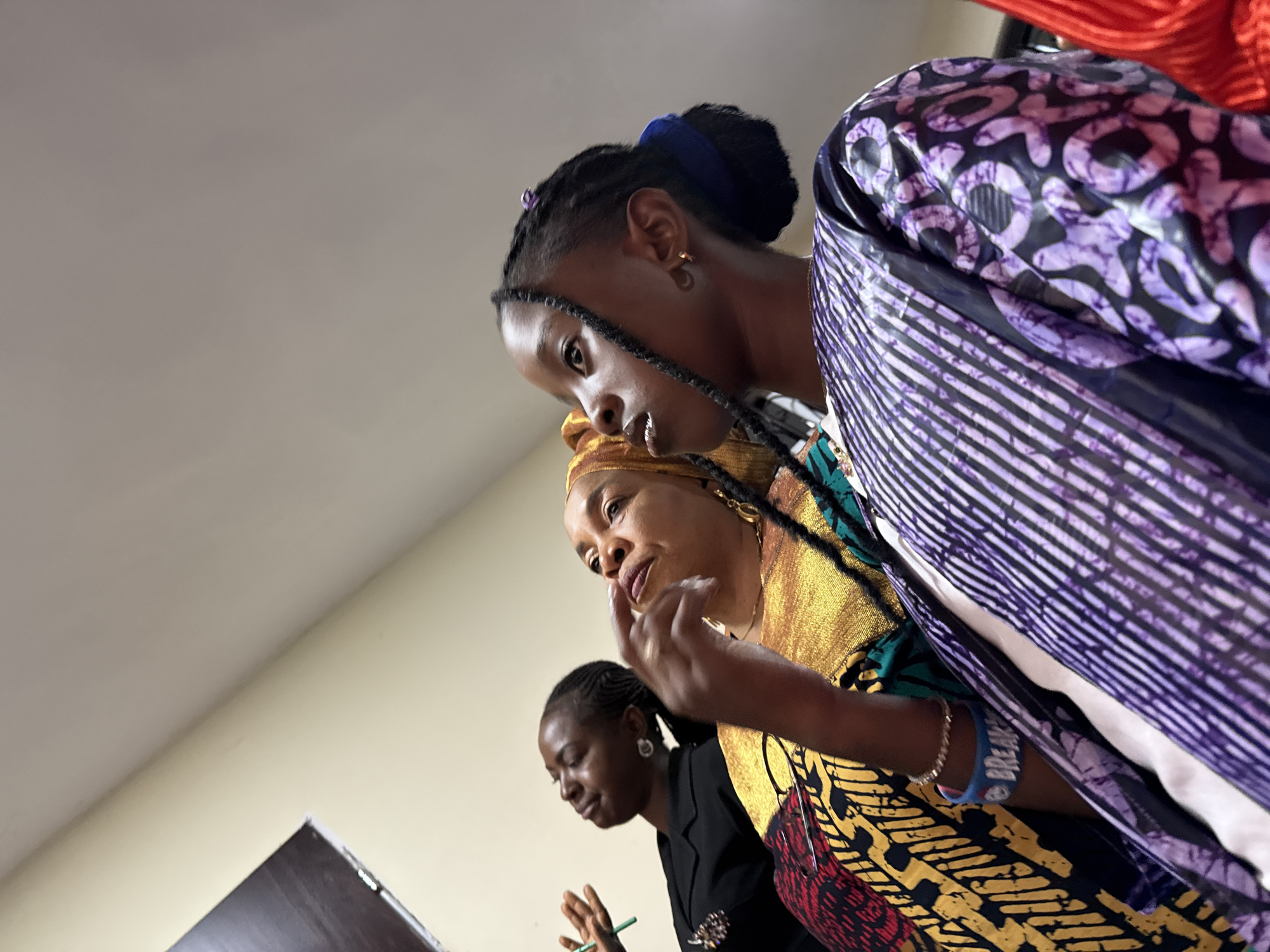 In Lugbe, Nigeria, an indoor scene depicts three women dressed in vibrant African attire. The central focus is on a woman in the foreground, wearing a gold and black patterned garment with a gold headscarf. She is looking down and to the left, with her right hand raised, index finger pointing as if speaking or emphasizing a point. To her left and slightly behind her, another woman wears a black blazer and appears to be looking downwards, possibly at a document or object. She holds a green pen in her right hand, with her fingers splayed. Further to the left and slightly out of focus, a third woman is partially visible. Dominating the right side of the frame is a woman wearing a purple and black patterned garment with a striped texture. The background consists of a light-colored wall and ceiling. The perspective is from a low angle, looking upwards towards the subjects.