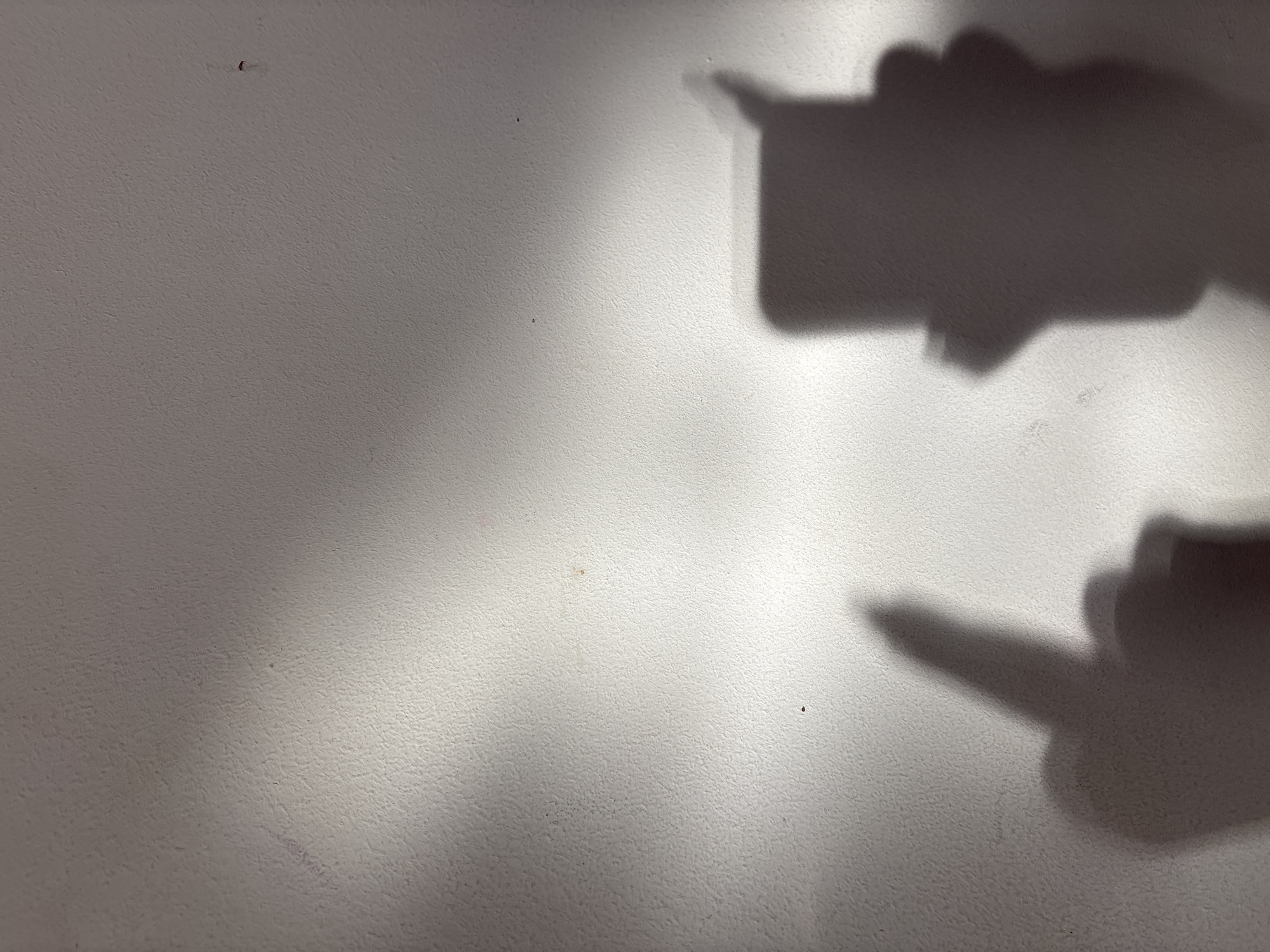 The image depicts a textured, off-white surface, likely a wall. A diffused, soft light source illuminates the wall from the bottom left, creating shadows on the right side. The shadows appear to be cast by hands and possibly an object like a smartphone. The shadows suggest a pointing gesture. There are some small blemishes or imperfections on the surface. The location cannot be determined from the image.