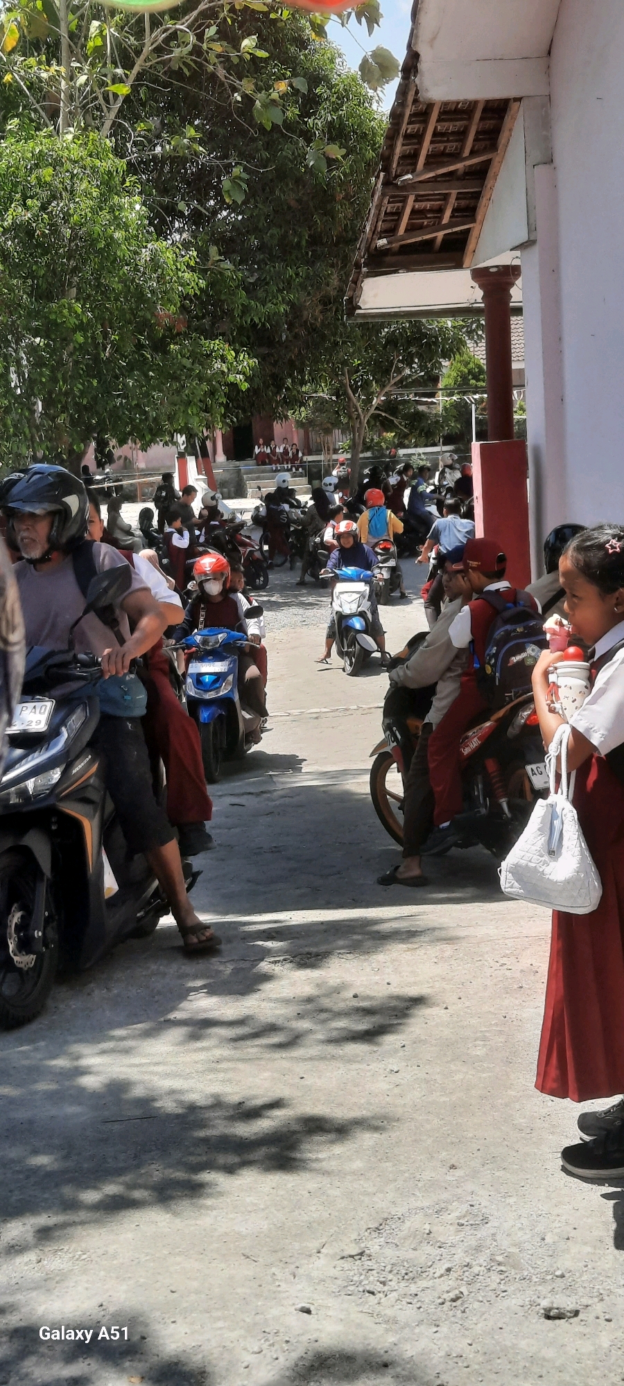 The image depicts a street scene in Tangkil, Indonesia, likely near a school, with numerous motorcycles and people. A group of students in red and white uniforms are visible in the background, some seated on a low wall or steps, while others are on motorcycles. Many individuals, including adults and children, are riding motorcycles, some wearing helmets. The environment appears to be outdoors with trees providing shade, and the ground is paved. The architecture of a building with a tiled roof and pillars is also visible. The overall atmosphere suggests a busy everyday scene, possibly during school dismissal or arrival.