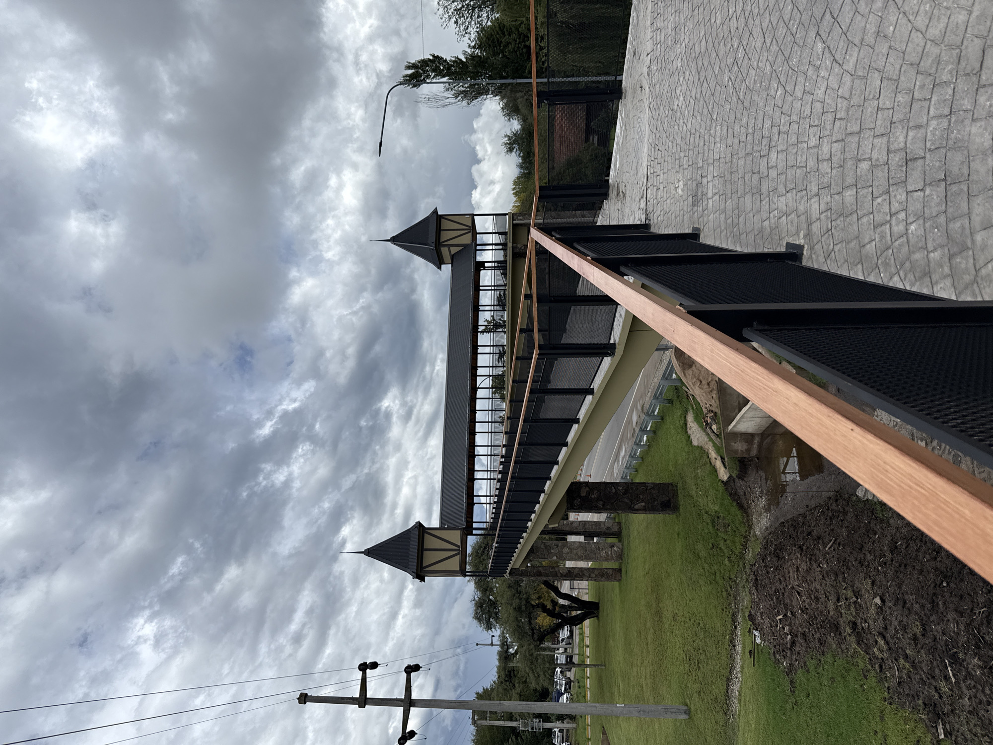 The image depicts a covered pedestrian bridge in Villa General Belgrano, Argentina. The bridge has two prominent towers with pointed roofs. The bridge structure is supported by stone-faced pillars. The bridge has black metal railings and a light-colored wooden handrail. There is a paved walkway on the top right and a grassy area with trees and parked cars visible below the bridge. There are utility poles with power lines in the foreground. The sky is overcast with grey clouds.