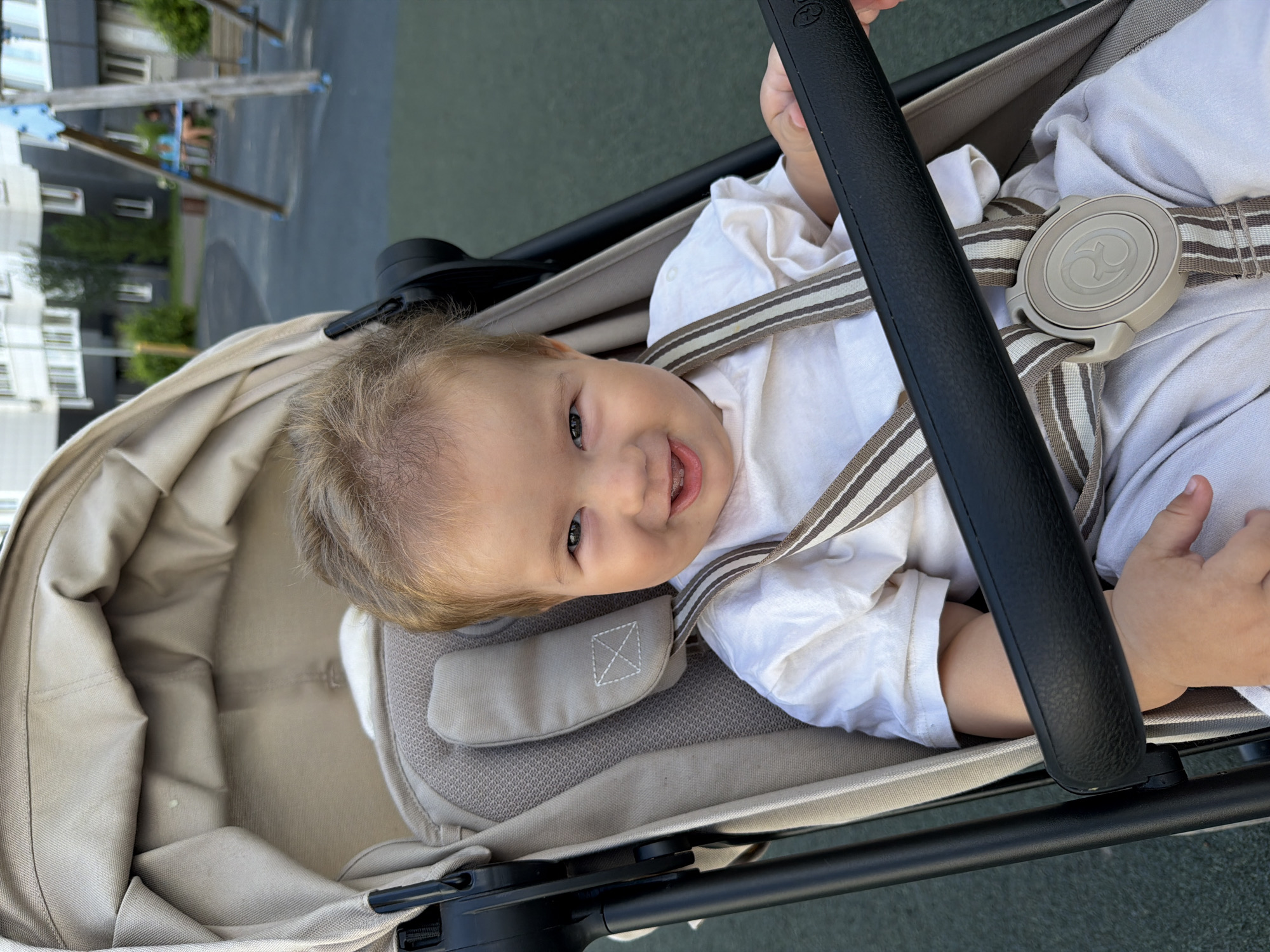 A child is in a stroller, smiling at the camera. The child has blonde hair and is wearing a white outfit. The stroller is beige with brown and white straps. In the background, there is a playground with swings and buildings. The scene is outdoors during the day.