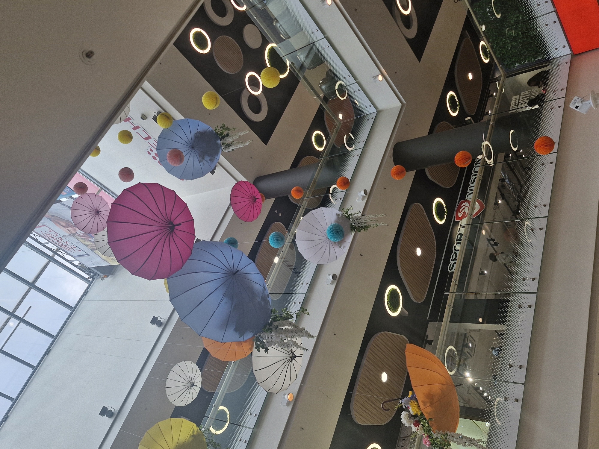 The image presents an upward perspective within the multi-story interior of a modern shopping mall, likely located in Sofia, Bulgaria. The primary focus is a vibrant decorative display of numerous colorful umbrellas suspended from the ceiling. Open umbrellas in shades of pink, light blue, orange, white, and yellow are visible, some adorned with floral arrangements or small decorative pom-poms in matching colors like yellow, orange, and light blue.
The mall's architecture features glass railings separating different levels, which reflect distorted images of the interior, including blurry figures of people, and additional ceiling details. The ceilings and walls are designed with geometric patterns, incorporating circular lighting fixtures, some of which frame green moss-like elements, and others simply providing illumination. A prominent dark, cylindrical structure, possibly a ventilation duct or a large architectural beam, spans horizontally across the upper part of the frame.
Through the reflections in the glass, the text "SPORT VISION" is clearly legible on a store sign, indicating one of the establishments within the mall. The overall scene conveys a cheerful and inviting atmosphere, characteristic of a contemporary retail environment, with the umbrella display adding a festive and whimsical touch. The lighting is artificial, suggesting the image was taken during operating hours indoors.