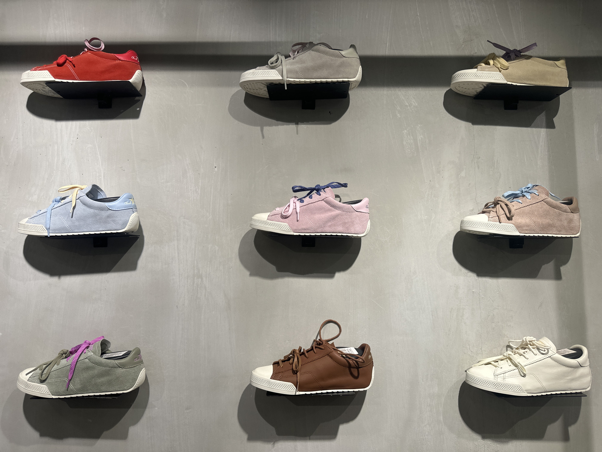 Nine shoes are displayed on a textured gray wall. The shoes are all low-cut sneakers with white rubber soles and colored suede or leather uppers. The colors include red, light gray, tan, light blue, pink, brown, and white. Each shoe is mounted on a small, black bracket attached to the wall. The scene appears to be a shoe store. The specific location within Kuta, Indonesia is not identifiable from the image.