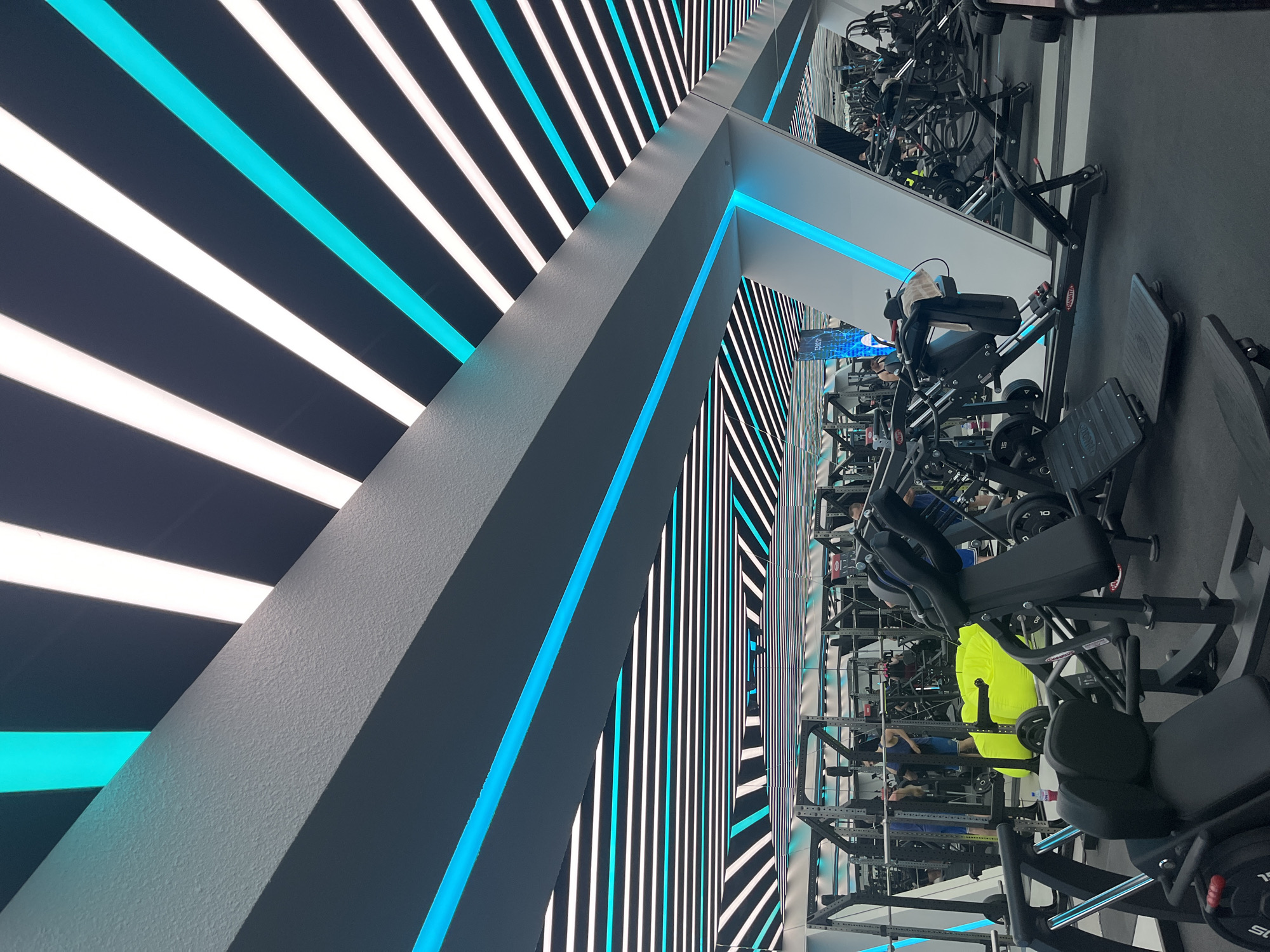 The image depicts the interior of a gym in an unknown city. The architecture features a striking design with alternating stripes of white and cyan light illuminating the ceiling and walls. A large, gray structural beam runs diagonally across the frame, accented with cyan lighting. The gym equipment, including leg press machines, is arranged in rows. Several people are using the equipment. The overall atmosphere is modern and well-lit.
