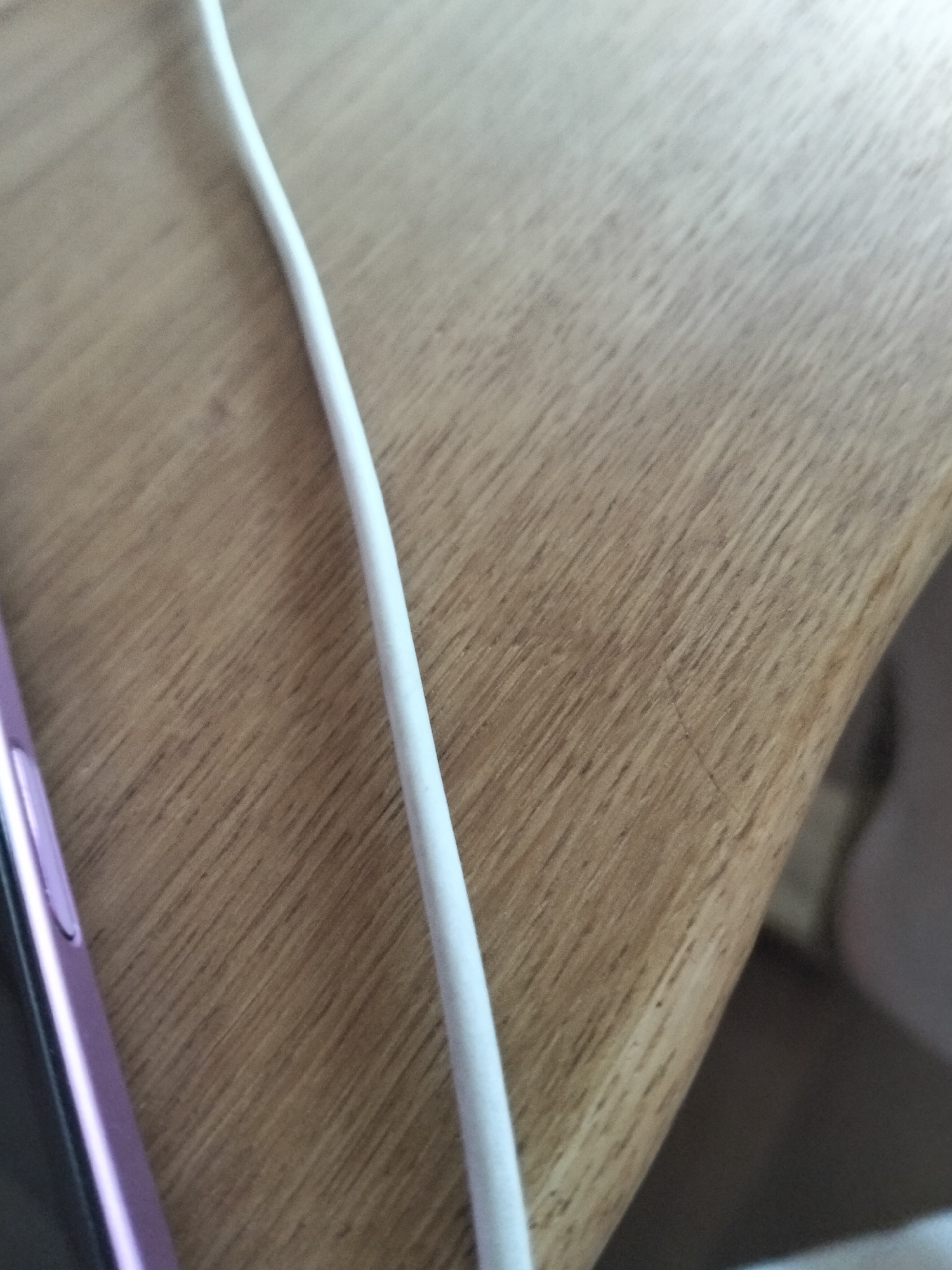 An extreme close-up shot shows a white cord running diagonally across a wooden surface. The wood grain is visible and runs diagonally in the opposite direction to the cord. On the left side of the image, a portion of a pink smartphone is visible. The shot is taken from above and to the side, with the lighting causing some glare on the cord and the phone. The location is not specified.