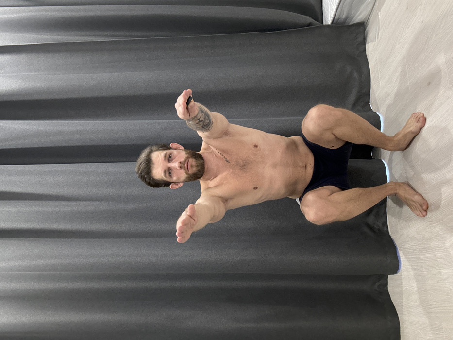 The image presents an adult male engaged in a bodyweight squat exercise within an indoor setting. The Caucasian male, with a beard and brown hair, is shirtless, wearing dark blue athletic shorts, and is barefoot on a light-colored wooden floor. His arms are extended forward at shoulder height, palms facing down, with a small, dark, rectangular object held in his right hand. A tattoo featuring a floral or abstract pattern is visible on his left forearm. He maintains a neutral expression, looking directly forward.
The background is dominated by a full-height, dark grey pleated curtain. A section of a white, textured object, consistent with a mattress or upholstered furniture, is partially visible on the far right, positioned behind the curtain. A faint blue light emanates from the base of the curtain on the right side.
The specific location within Saint Petersburg, Russia, cannot be determined from the visual information provided in the image.