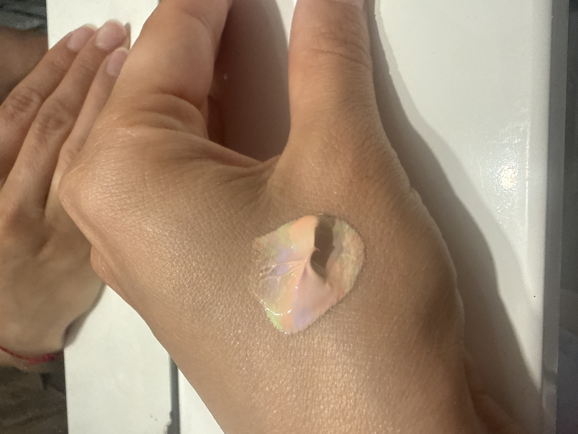 A light-skinned hand with short, unpainted fingernails has a dollop of multicolored cosmetic product on the back of the hand. The product contains colors of peach, green, and grey. The hand is set against a white surface, which may be a table. Another hand, and potentially arm, appears in the upper left corner, near the edge of the frame. The location is not identifiable from the image.