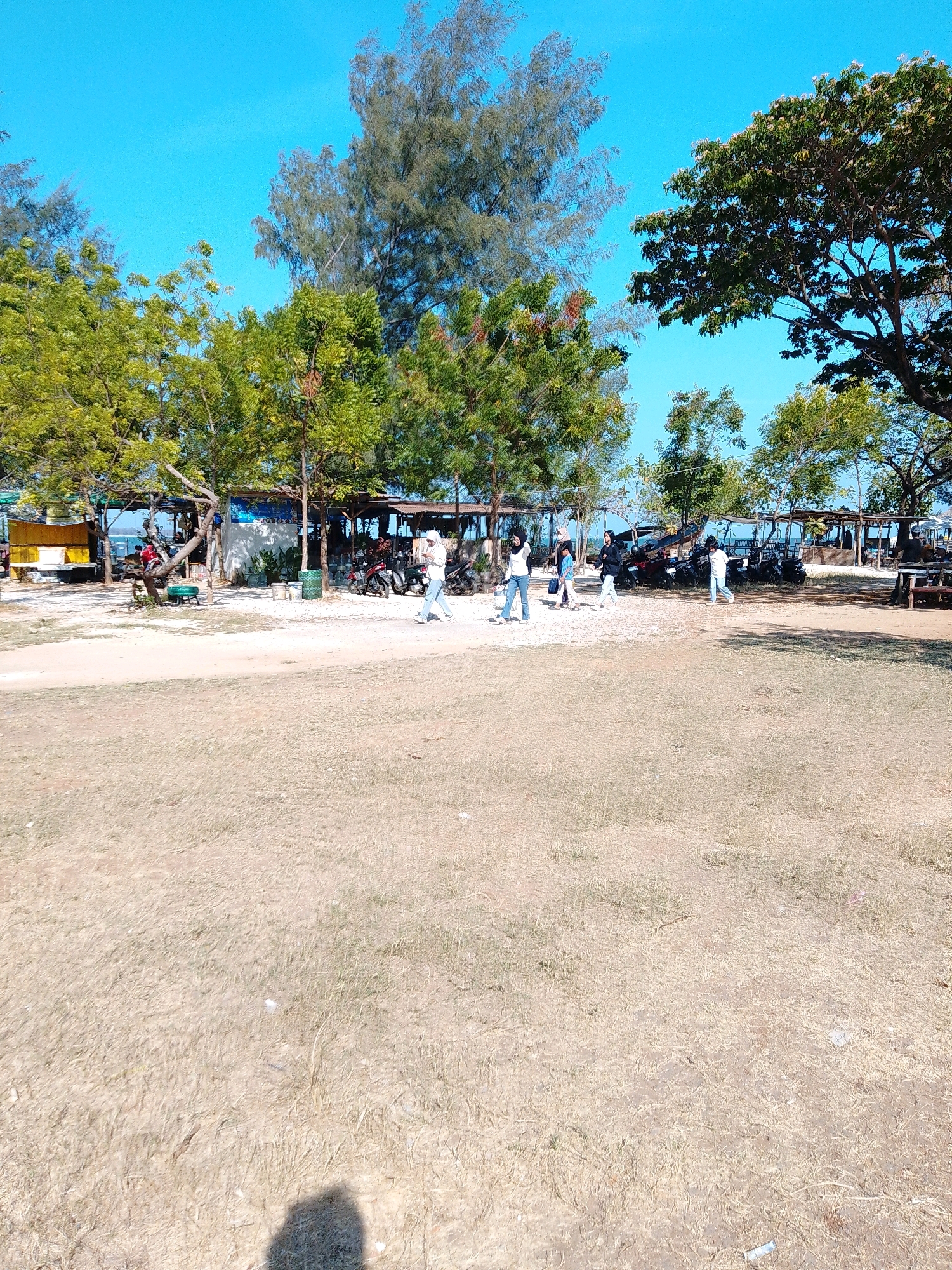 The image depicts a coastal area in Cikal, Indonesia, under a clear blue sky. The foreground is dominated by dry, sparse vegetation on sandy ground, with a shadow of a person visible in the lower right corner. In the midground, several trees with green foliage line a sandy path. Behind the trees, a row of simple open-air structures, likely stalls or shelters, is visible. Several motorcycles are parked in front of these structures. A few people are walking on the sandy area in front of the stalls. In the background, the ocean can be seen, with a bright horizon line. The overall impression is of a casual, perhaps tourist-oriented, beachside location.