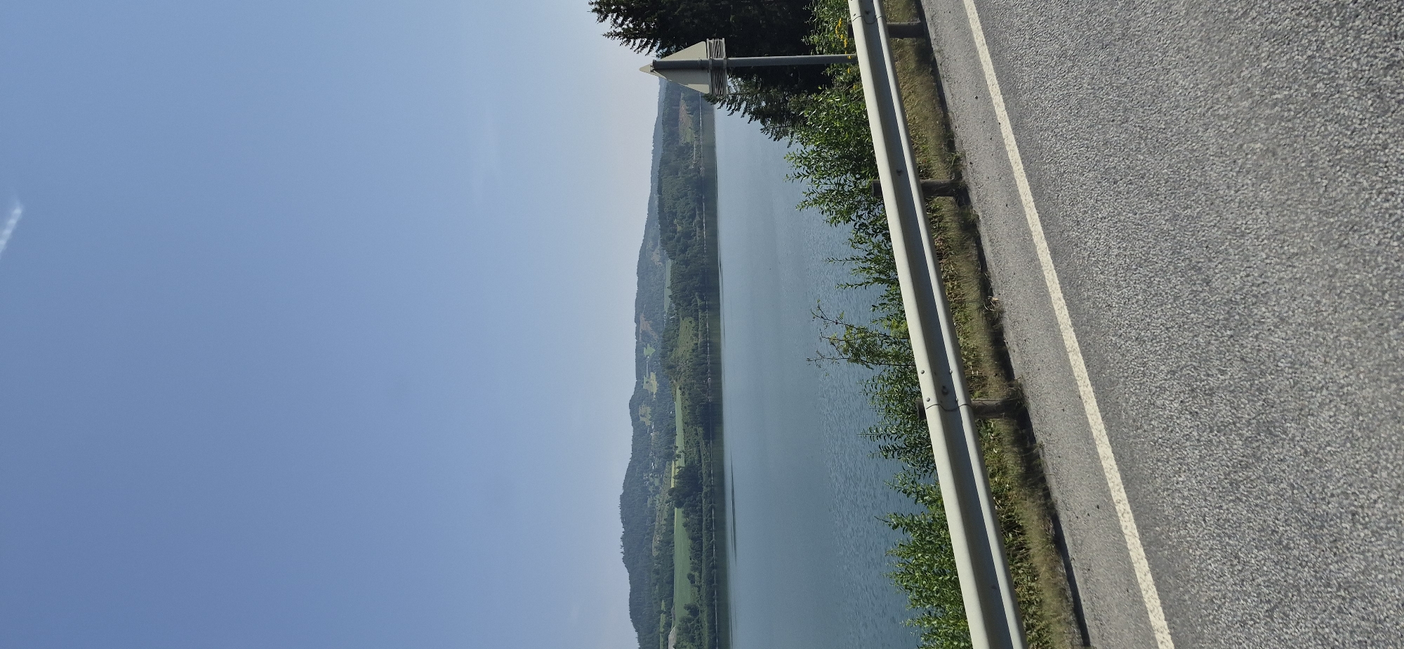 The image shows a scenic view of a body of water with rolling hills in the background. In the foreground, there is a road with a guardrail and some vegetation. The sky is clear and blue. There are no people or specific landmarks visible that would indicate the location in Vingrom, Norway. The scene appears to be a natural landscape with infrastructure elements.