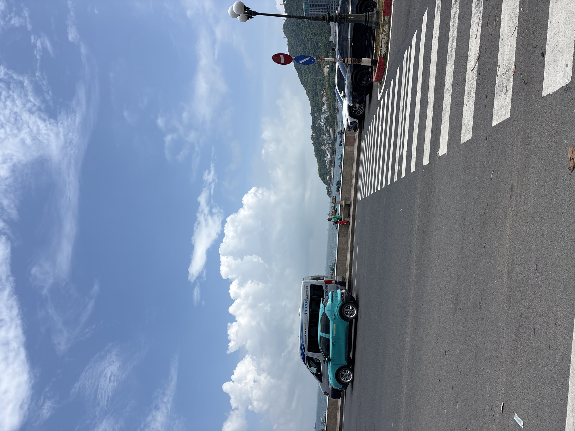 The image depicts a street scene with a pedestrian crossing in the foreground. A bright turquoise car is parked on the left side of the street, beside a silver van with "AN PHU EXPRESS" written on its side. In the background, there are several vehicles, including a white SUV and a black SUV, parked near a scenic coastline with a hill covered in buildings and trees. A lamppost with multiple lights stands to the right of the black SUV. The sky is blue with scattered white clouds. There is a person in a green shirt sitting on a bench on the left side of the image, facing the sea. The overall scene suggests a coastal city or town, likely in Southeast Asia, given the architecture and the type of vehicles. This scene is not specifically identifiable as being within Ho Chi Minh City, Vietnam, but it is consistent with urban coastal areas of Vietnam.