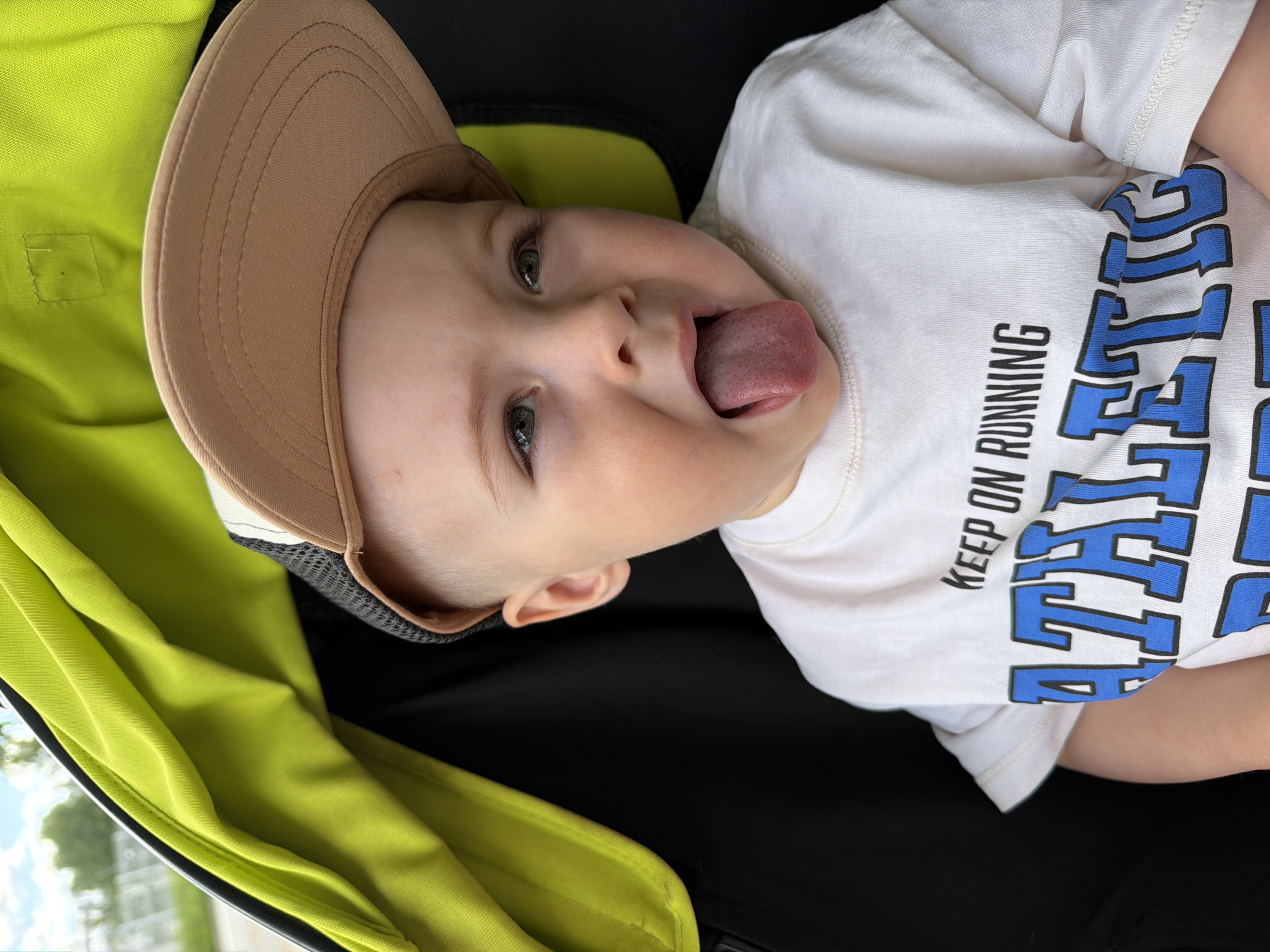 A close-up shot shows a young child in a stroller. The child is wearing a white t-shirt with blue and black lettering that reads "ATHLETIC" and "KEEP ON RUNNING." They are also wearing a tan baseball cap with a black mesh back. The child is sticking their tongue out. The stroller has a bright lime green interior lining. The background is blurred, suggesting an outdoor setting with some greenery and a cloudy sky. The image was taken in Votkinsk, Russia.