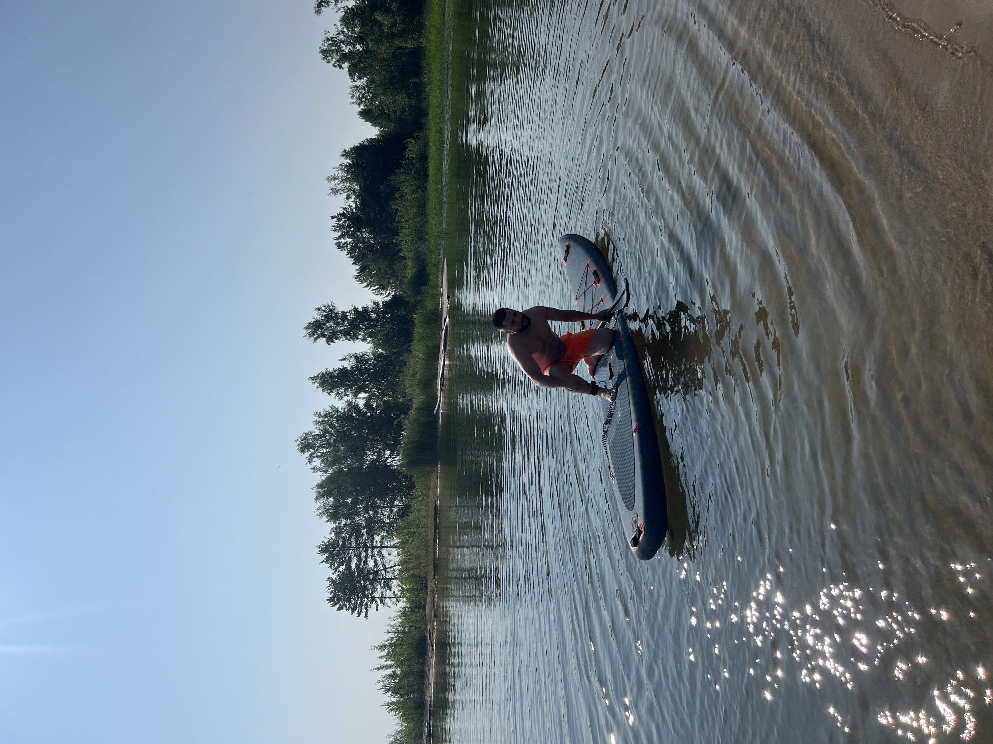 An adult male, with a beard and dark hair, wearing orange swim trunks, kneels on a stand-up paddleboard in a body of water. The paddleboard is dark gray with red accents and has a paddle lying on it. The man is positioned in the center of the frame. The water is calm with ripples and sun reflections on the surface. In the background, a sandy shore is visible, leading to a line of trees that forms the horizon. The sky is clear and blue, with the sun creating a lens flare in the upper left corner. The scene appears to be a natural outdoor environment, likely a lake or river. The location is identified as the city of Borovoe, Russia.