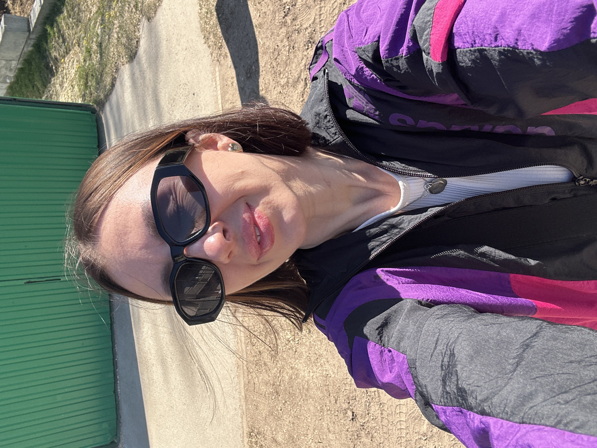 A young woman wearing black sunglasses, a white ribbed top, and a purple, black, and pink windbreaker stands outside on a sunny day. Behind her are a green metal fence, a concrete curb, a sidewalk, and an area of dry grass or dirt. The location is undetermined; reference to the city of Русский Вожой, Russia is unsupported.
