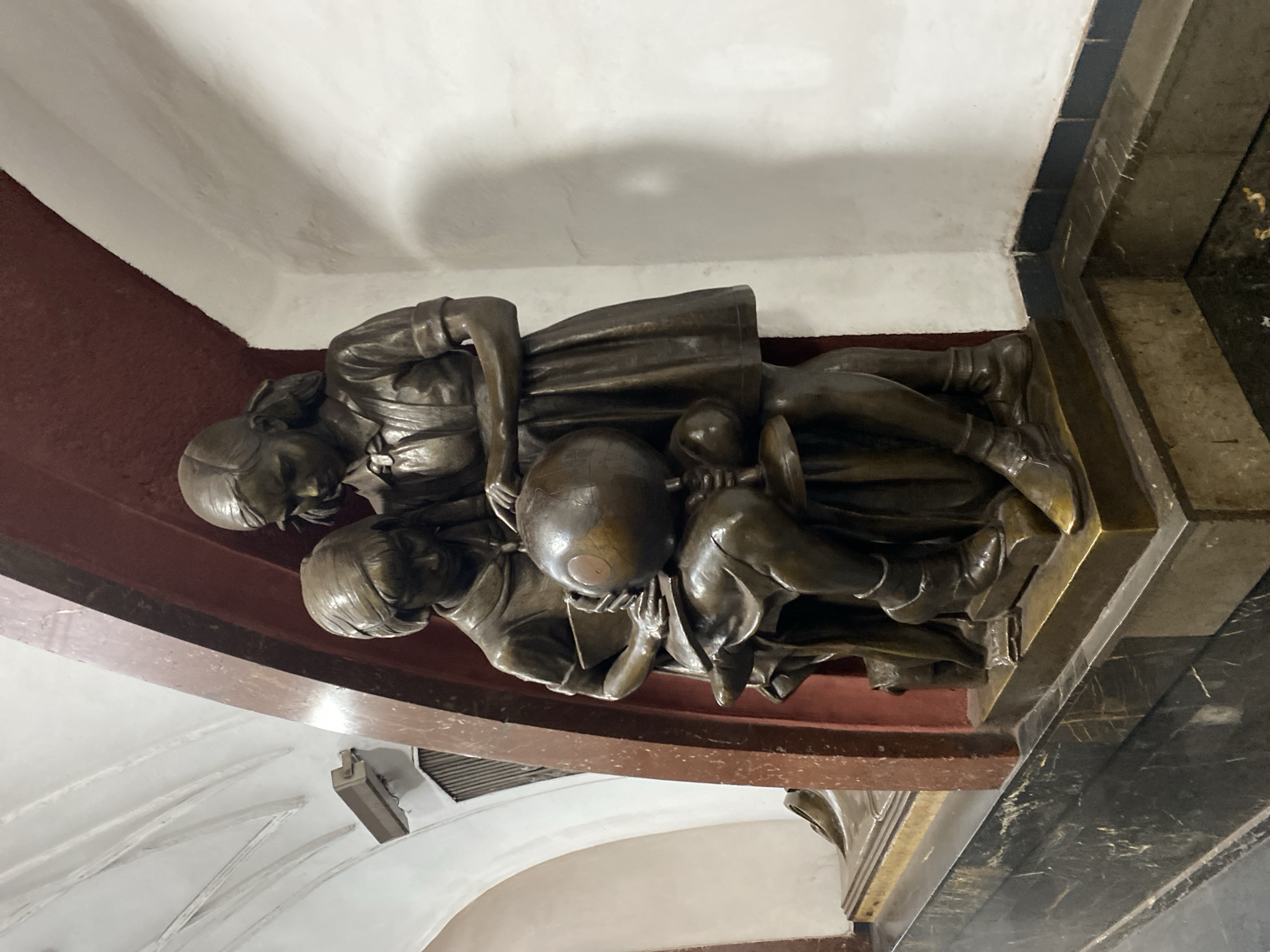A bronze bas-relief depicts three figures, possibly children, huddled around a spherical object, likely a globe. They appear to be reading or studying, given their postures and focus on the globe. The sculpture is attached to a wall surface. The environment appears to be the foyer of the Kiyevskaya Metro Station in Moscow, Russia. The station is known for its ornate decorations, including mosaics and other sculptural elements. The red and white ceiling and the marble pillar visible in the background are architectural details of the Kiyevskaya station.