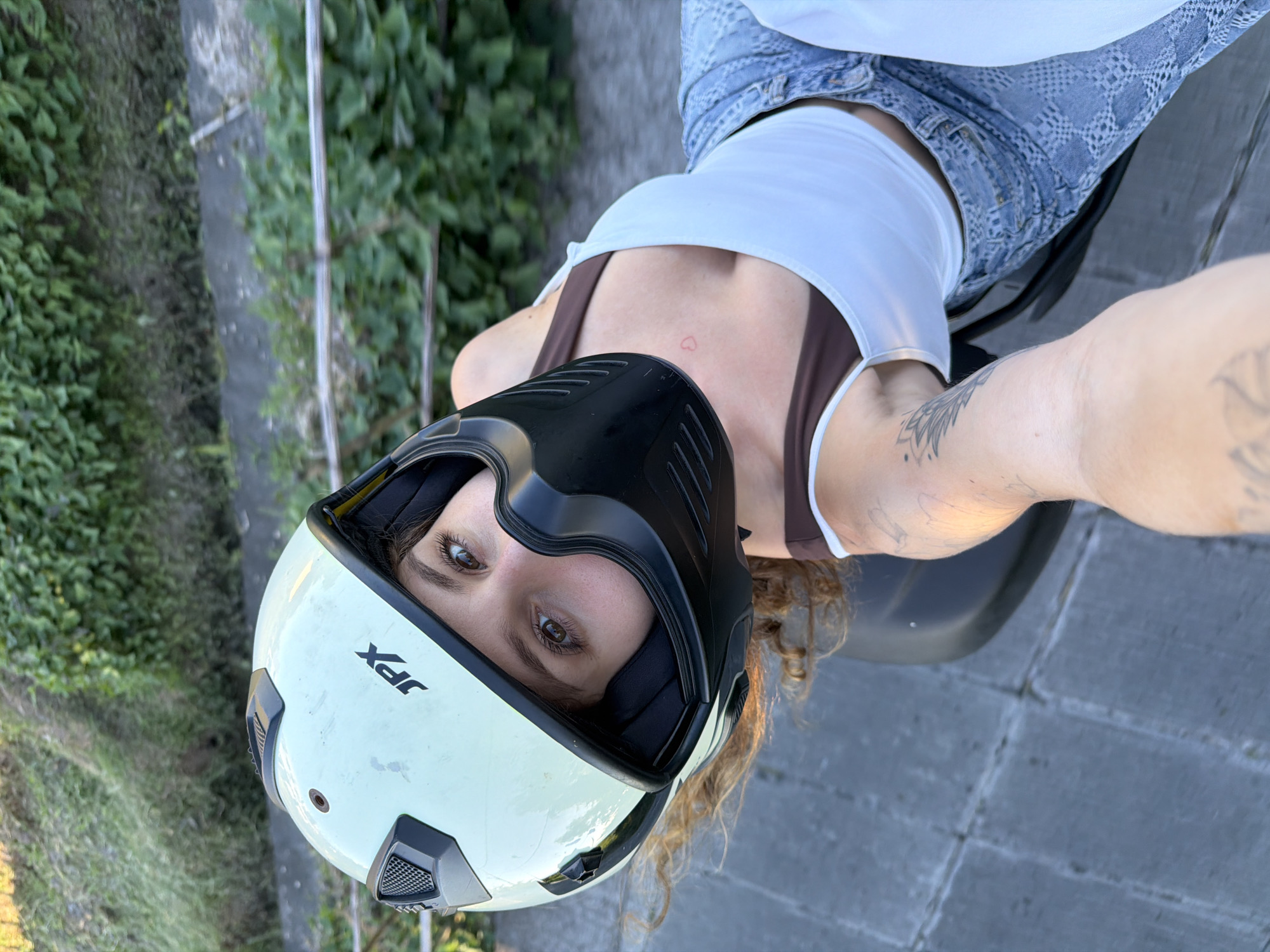 A young woman, wearing a white and black motorcycle helmet marked "JPX", is posing for a selfie while sitting on a motorcycle. She is wearing a brown and white tank top and denim shorts. She has tattoos on her left arm and a small heart tattoo on her chest. The background includes foliage and a gray paved area, possibly a sidewalk. The location could be North Kuta, Indonesia, based on the prompt.
