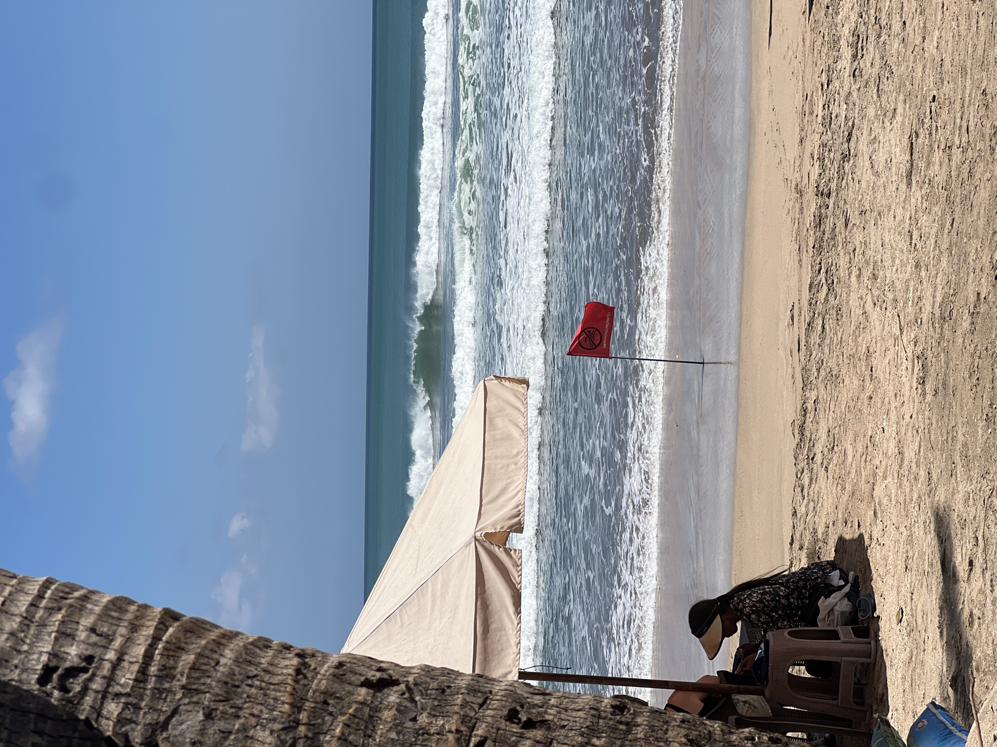 In Kuta, Indonesia, a beach scene unfolds under a clear blue sky with scattered white clouds. The foreground is dominated by the rough bark of a palm tree on the left, partially obscuring a beige beach umbrella. Beneath the umbrella, a person wearing a wide-brimmed hat and a floral-patterned shirt sits hunched over, facing away from the viewer. To their right, a small plastic stool is visible.
Further out, the ocean displays moderate waves with white foam, rolling towards the sandy shore. A single red flag, indicating "swimming prohibited," is planted in the sand near the water's edge. The beach itself is a light brown sand, showing some textured footprints and minor debris. The horizon is a sharp line between the deep blue of the sea and the lighter blue of the sky. The overall atmosphere is one of a sunny day at the beach, with the red flag suggesting caution for swimmers.