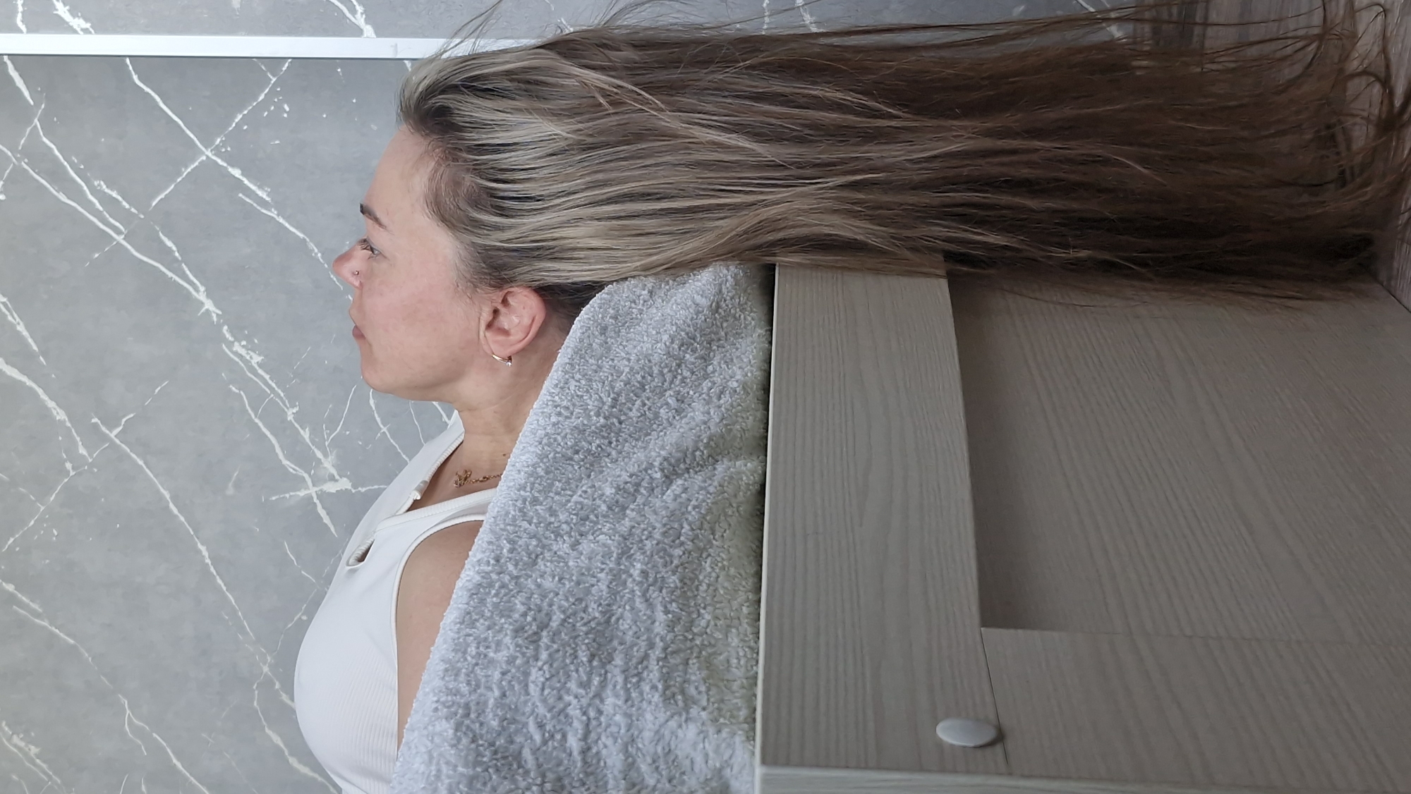 A woman with blonde and brown hair is lying down with her head on a gray towel next to a wooden surface. She is wearing a white tank top and a small gold necklace. She has a gold hoop earring in her left ear and a piercing in her nose. The background is a gray wall with white marble-like patterns. The location is not identifiable from the image.