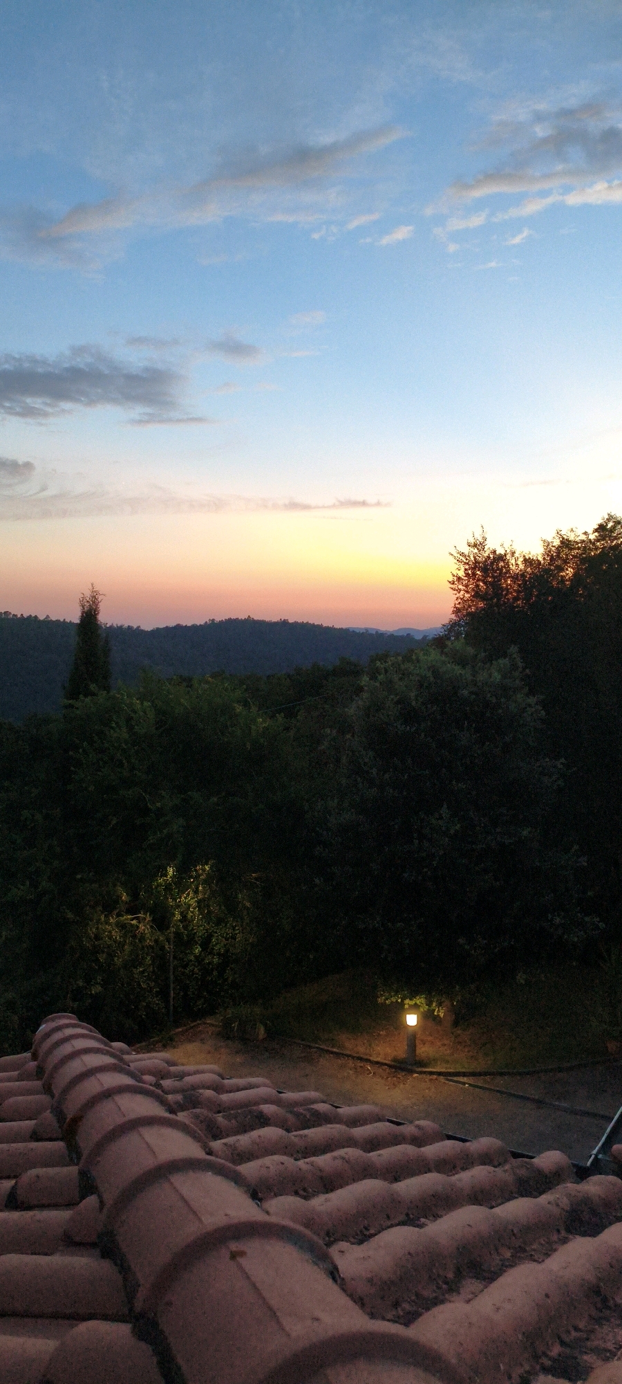 A vertical, medium shot captures a serene twilight landscape from a rooftop vantage point in Casale di Pari, Italy. The foreground is dominated by a close-up view of weathered, terracotta roof tiles, their rounded shapes and reddish-brown hue contrasting with the fading light. Beyond the roof, a dense, dark green forest stretches across the midground, its foliage appearing lush and abundant. A single, slender evergreen tree stands out against the darker canopy on the left. In the distance, rolling hills are silhouetted against a gradient sky transitioning from pale blue at the top to soft hues of pink, orange, and yellow near the horizon, indicative of sunset. A small, illuminated lamp post with a warm, yellow light is visible amidst the trees in the midground, casting a subtle glow on the surrounding vegetation. The overall atmosphere is one of quiet tranquility and natural beauty at the close of day.