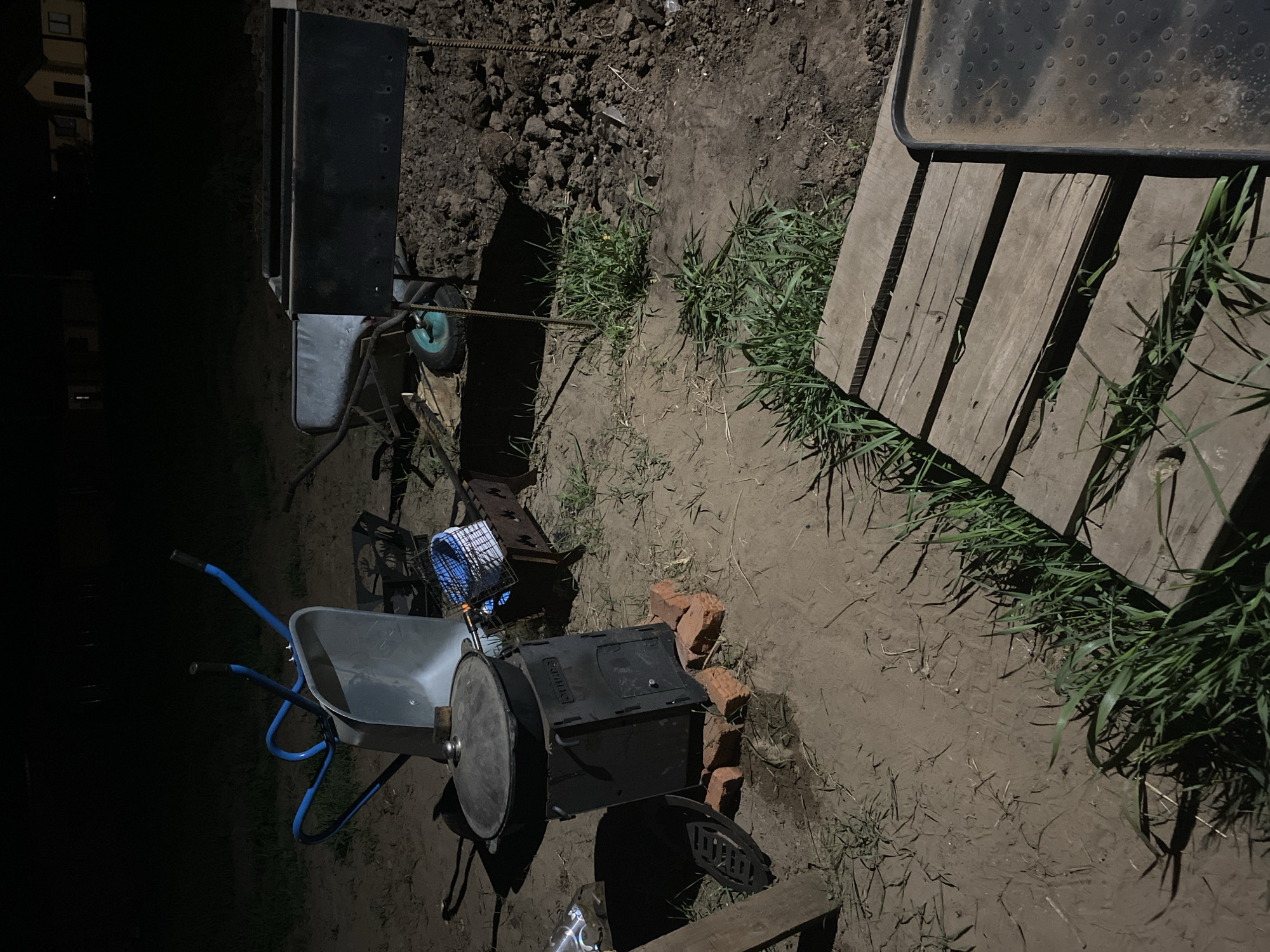 The image depicts an outdoor scene in the city of Unknown, Unknown, at night. The ground is dirt. Several objects are present including a wheelbarrow, a grill placed on top of bricks, wooden planks, and various pieces of metal possibly used for grilling. The lighting appears to be low or from artificial sources.